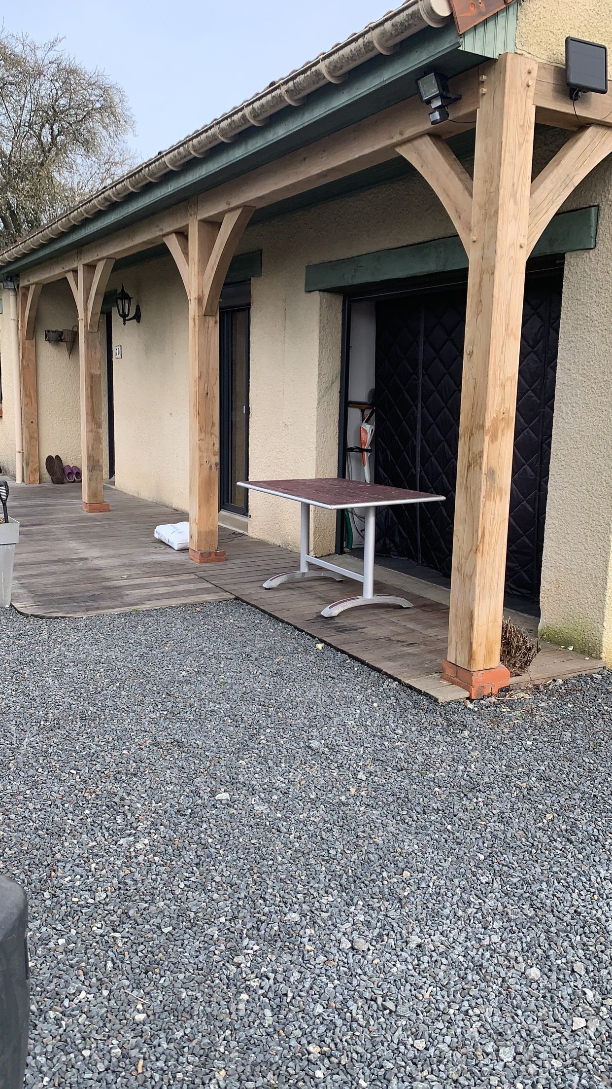 Structure à poutres en bois au-dessus d'une terrasse avec une table et un sol en gravier.