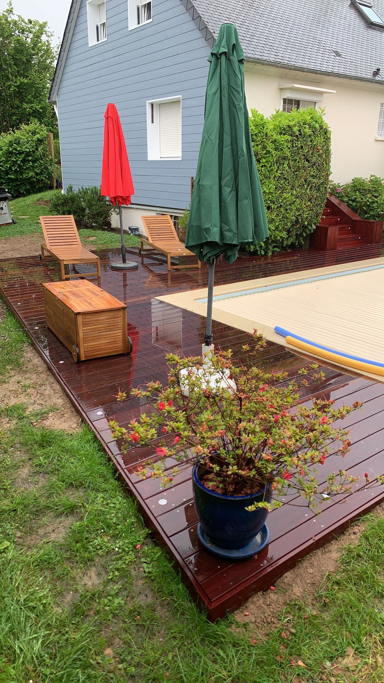Jardin avec terrasse rouge, piscine, chaises longues, parasol vert fermé, parasol rouge et pot de fleurs.