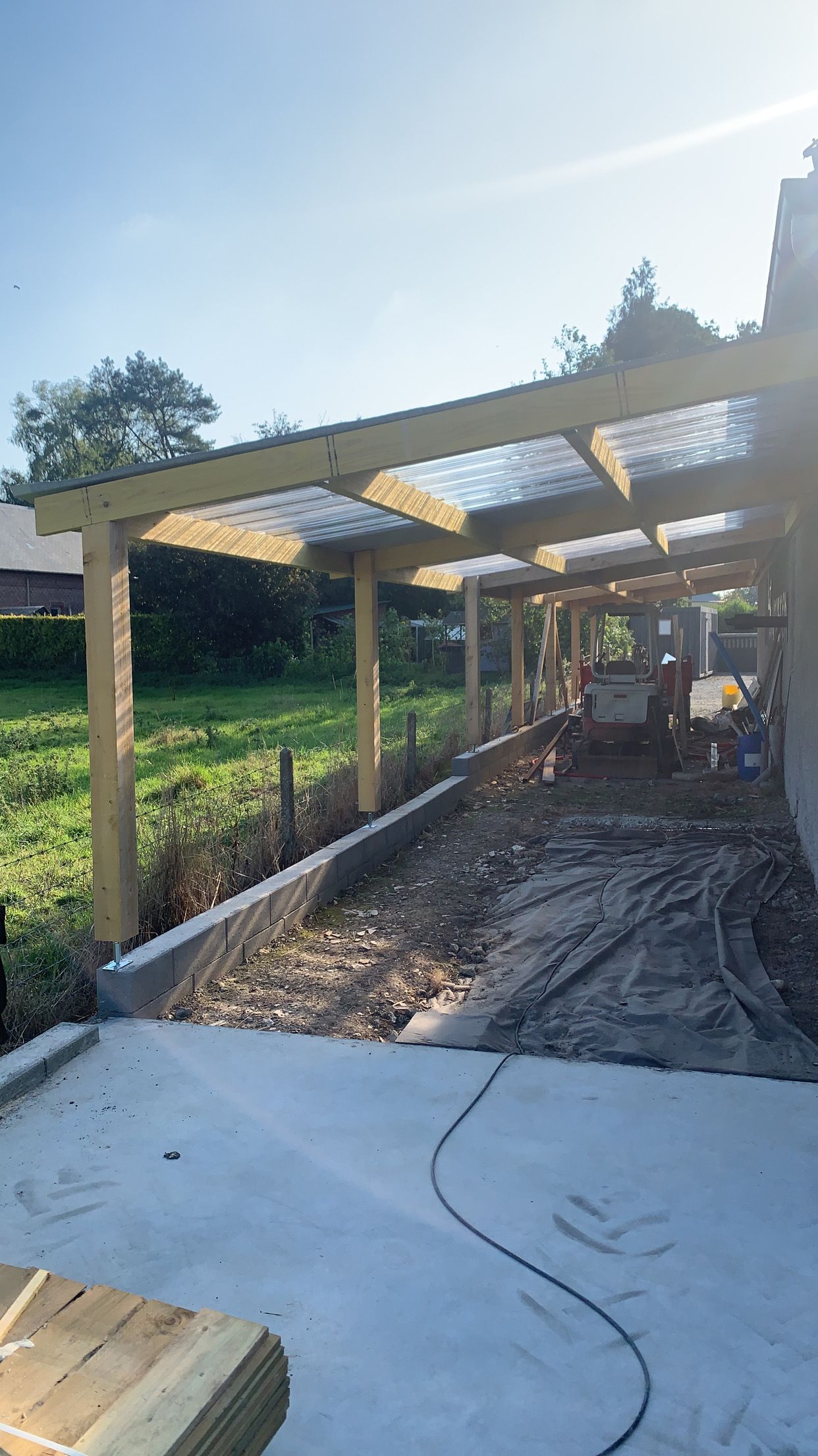 Pergola en bois en construction ; toiture translucide, base en béton et végétation environnante.