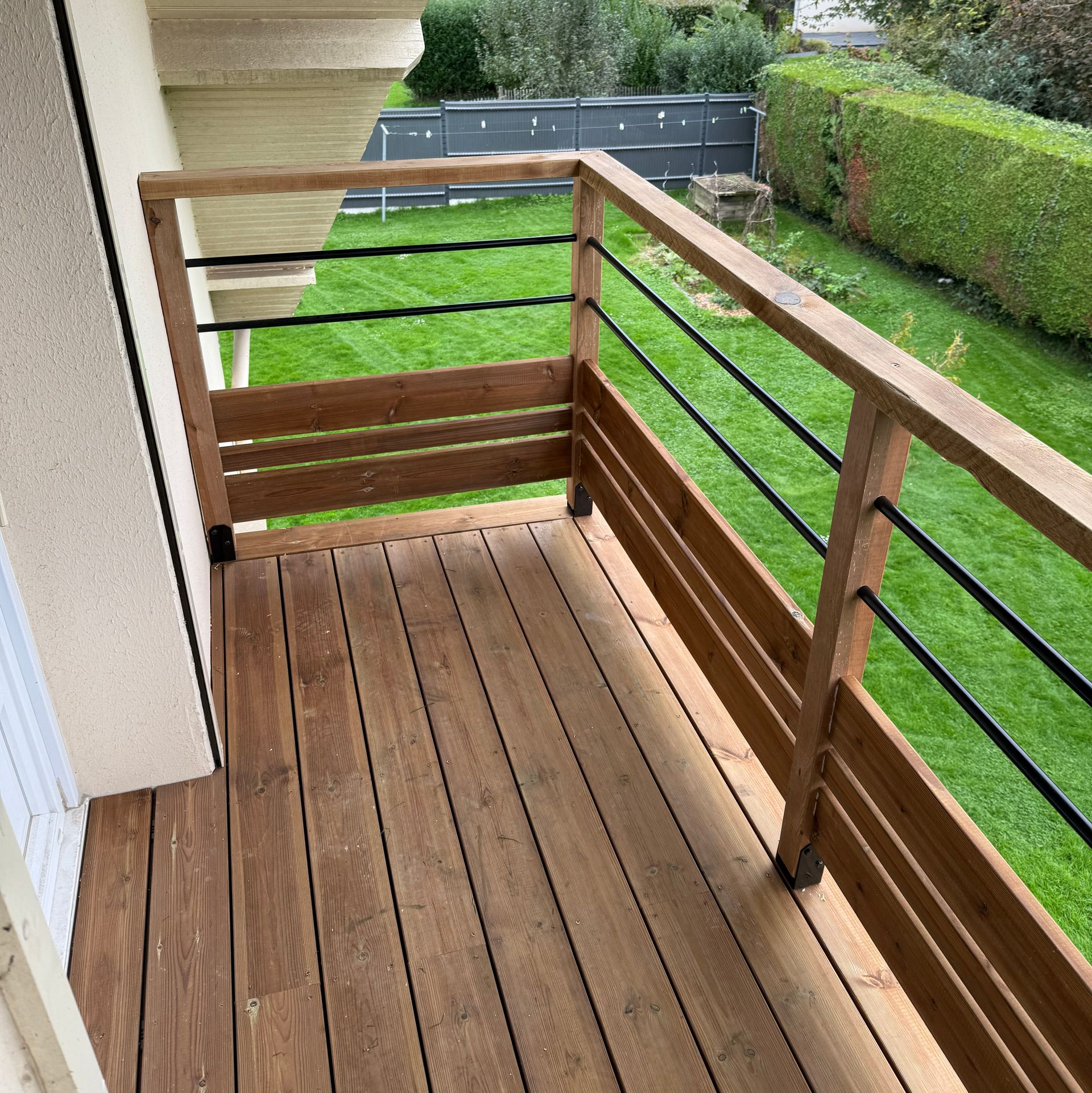 Petit balcon en bois avec une rambarde en bois et des barreaux en métal noir, donnant sur une cour gazonnée.
