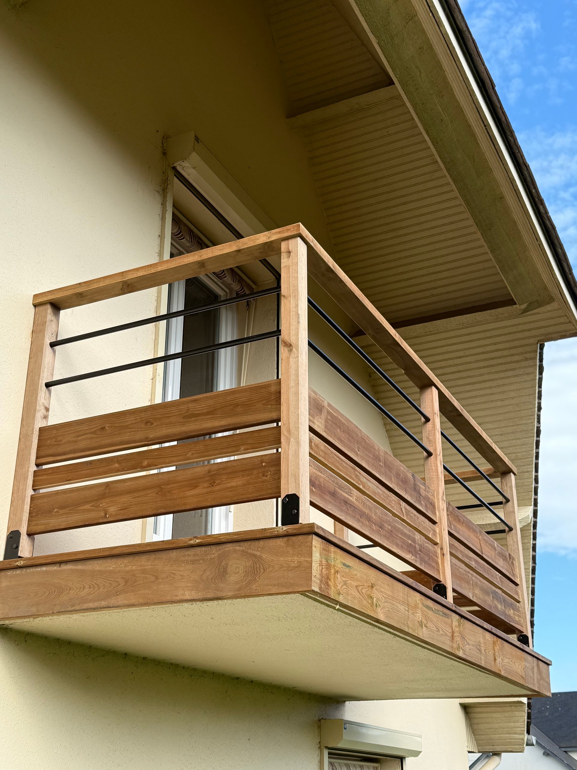 Balcon en bois à lattes horizontales, rambardes noires, attenant à un bâtiment beige, ciel bleu en arrière-plan.