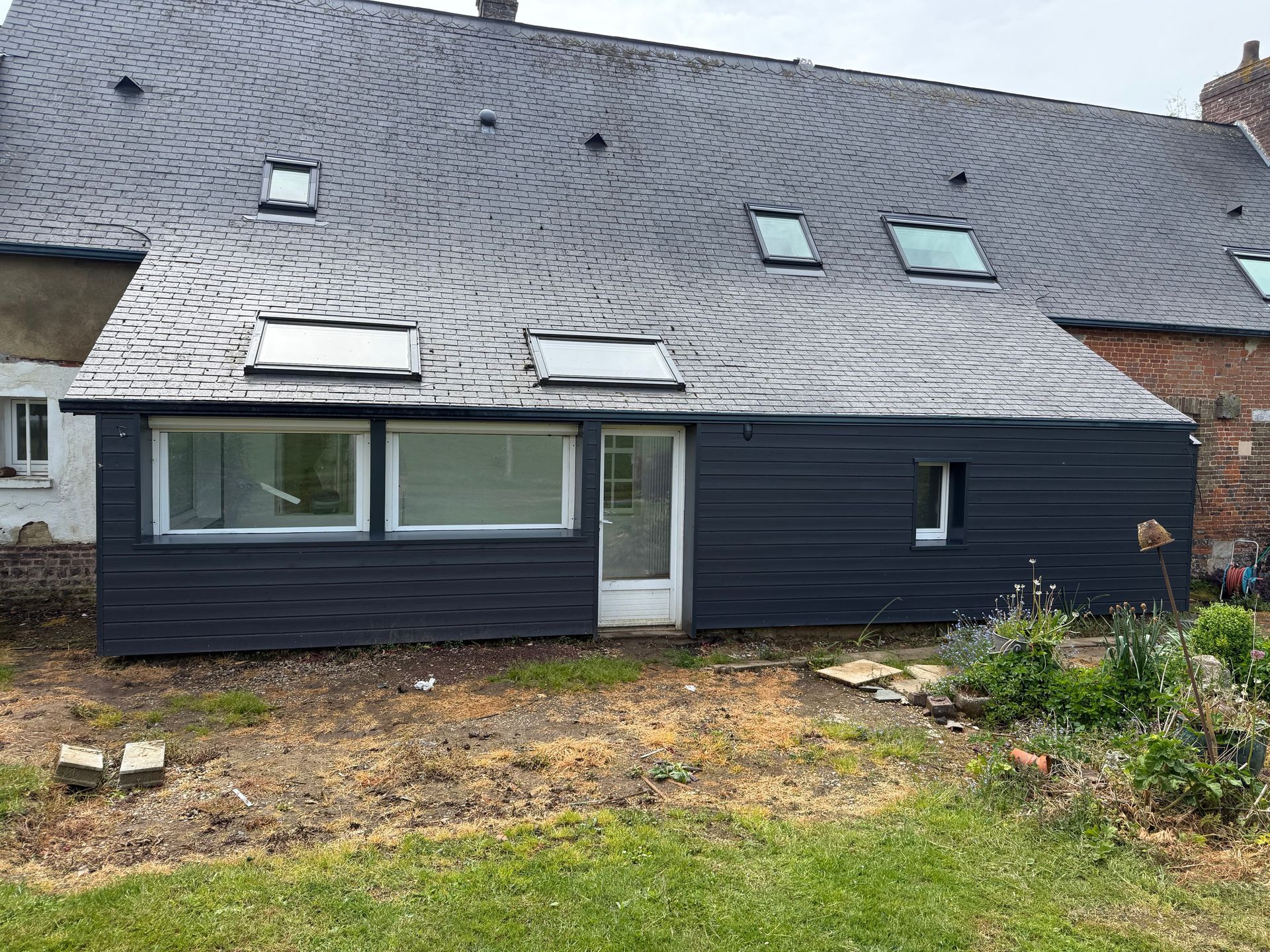 Bâtiment aux façades bleu foncé et percé de plusieurs puits de lumière, jouxtant un bâtiment délabré au toit d'ardoise.