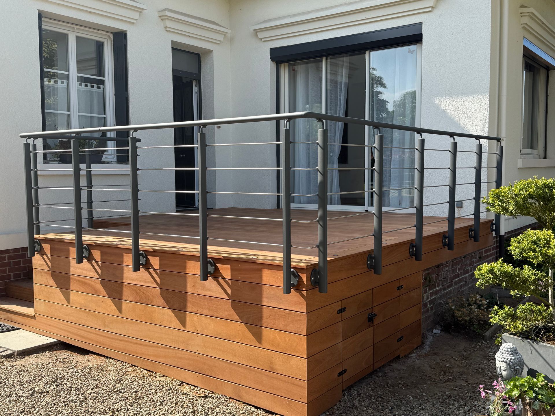 Terrasse en bois avec rambarde en métal noir, jouxtant une maison avec fenêtres.