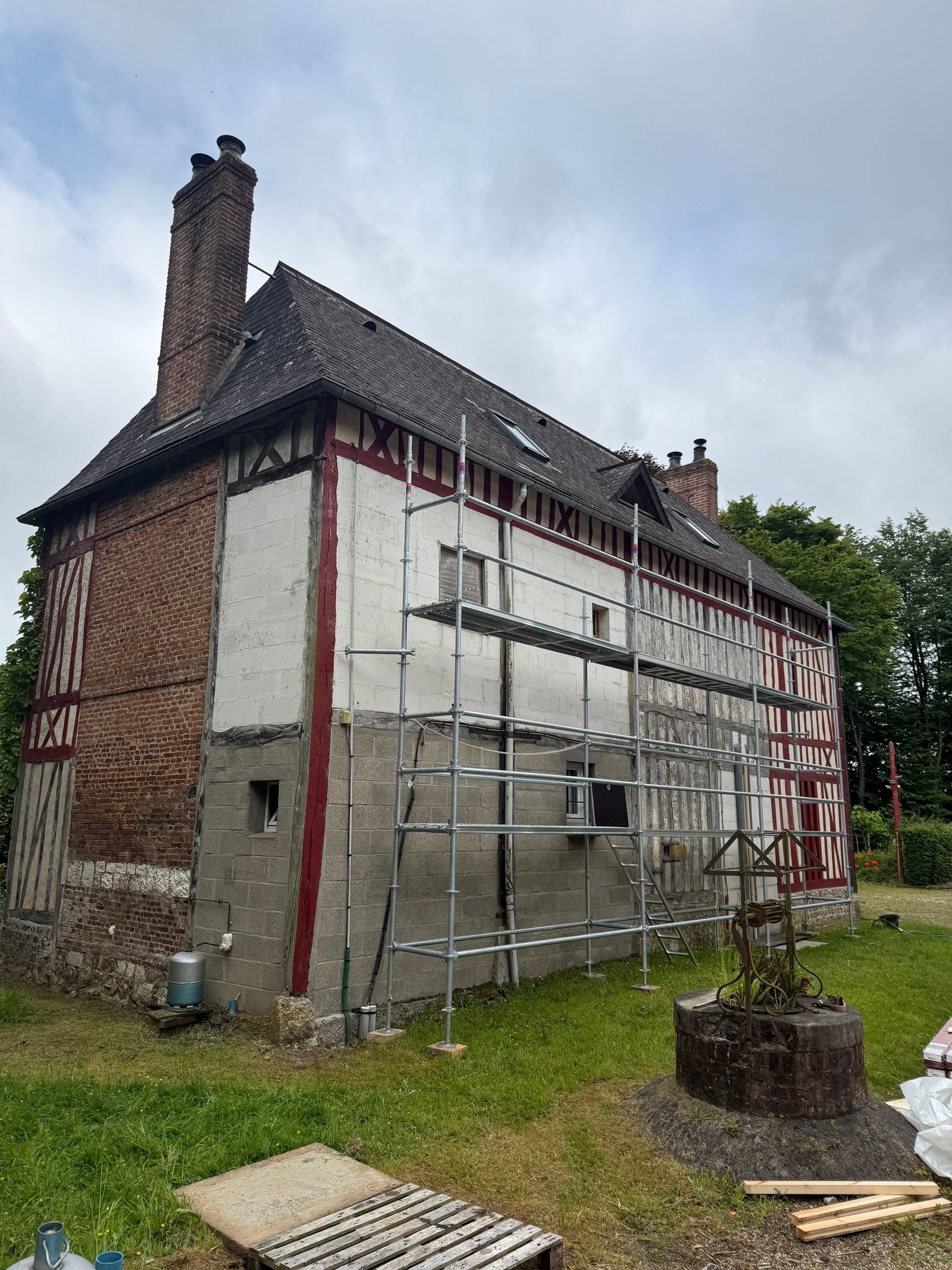 Bâtiment à colombages en cours de rénovation, avec échafaudages. Façade rouge et blanche, cheminée en briques, fondations en pierre.