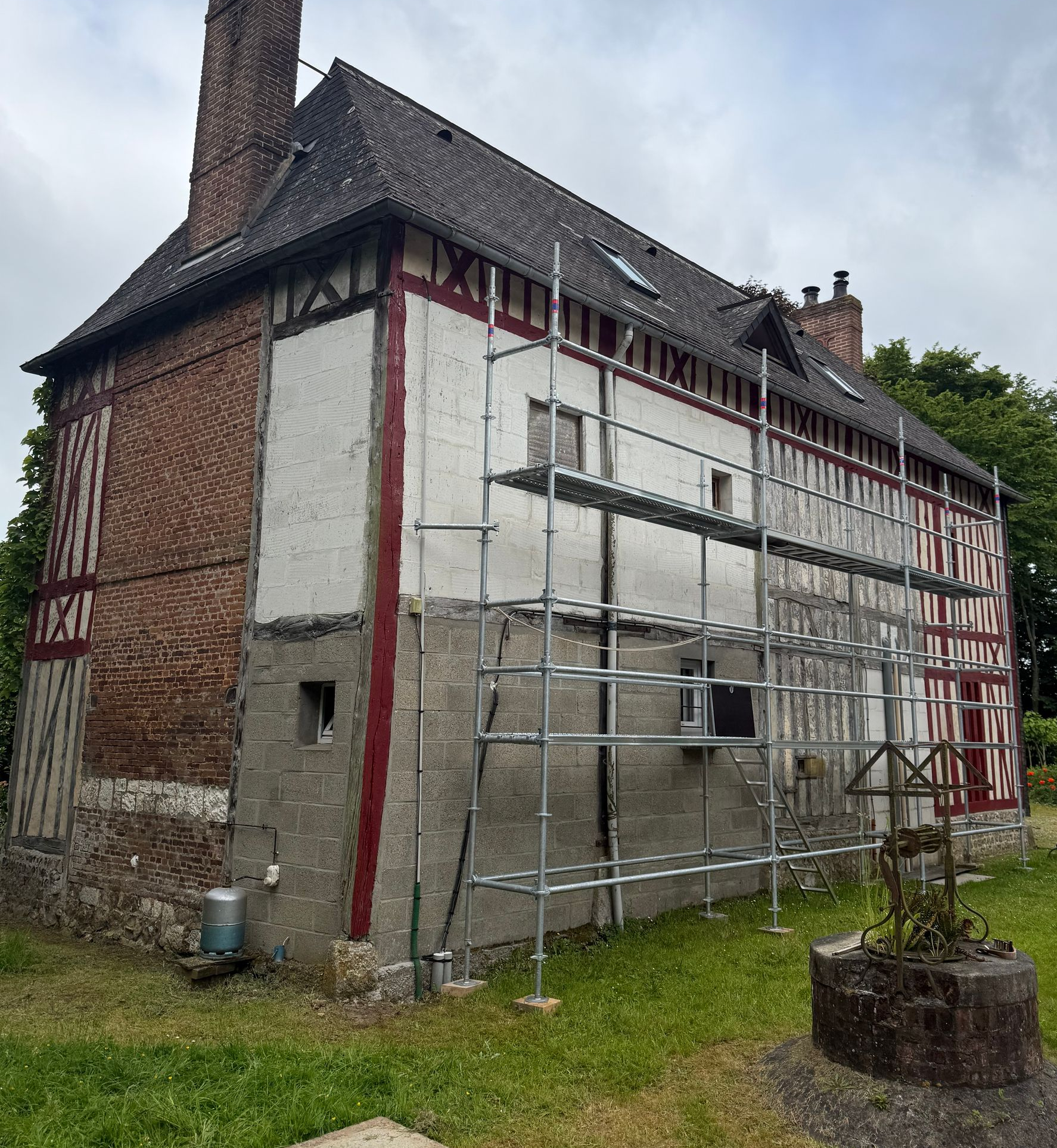 Maison à colombages en rénovation, avec échafaudages, boiseries rouges et cheminée en briques, sur une pelouse.