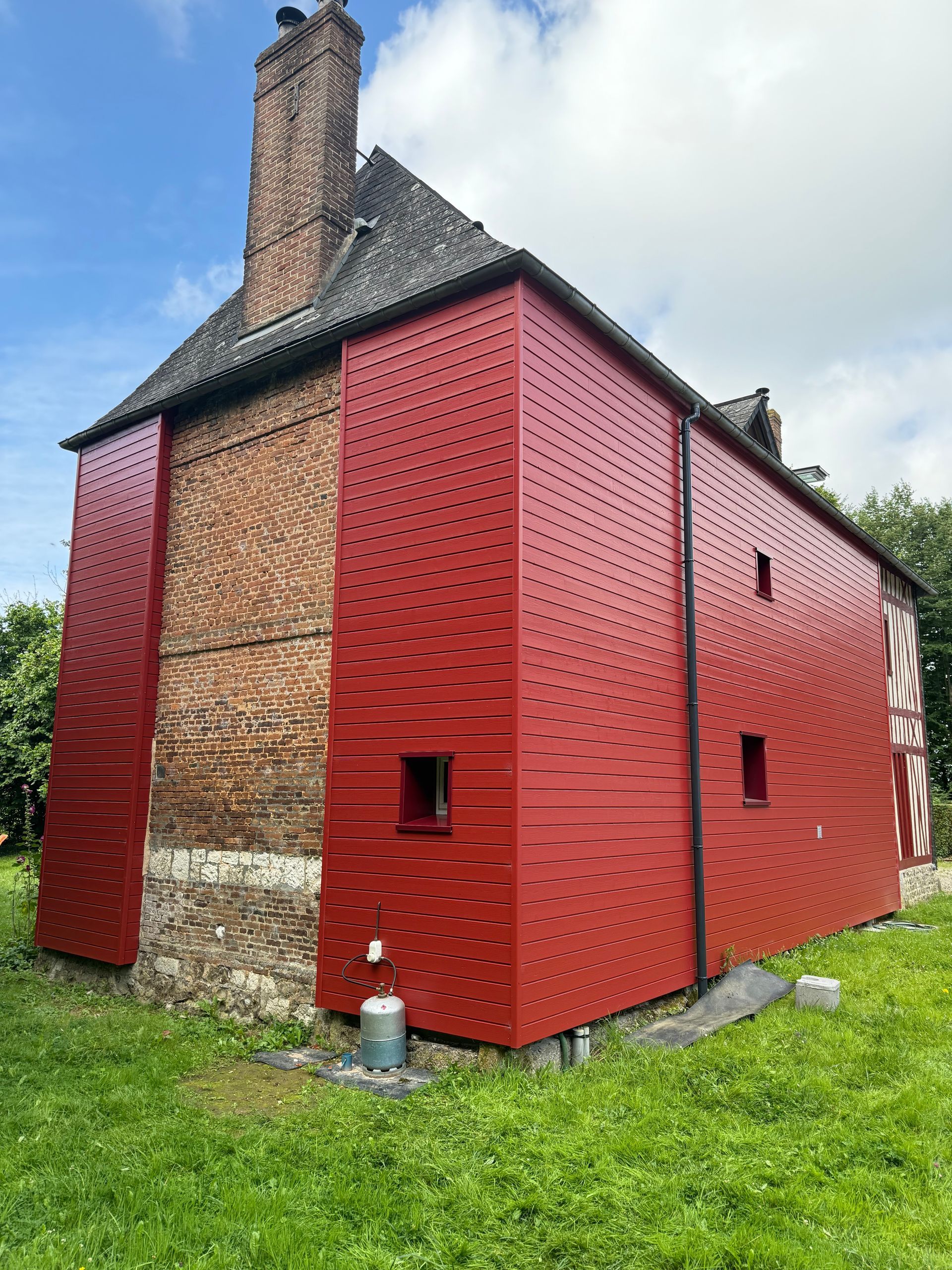 Bâtiment en briques recouvert d'un bardage métallique rouge perforé. Cheminée, soubassement en pierre et pelouse.