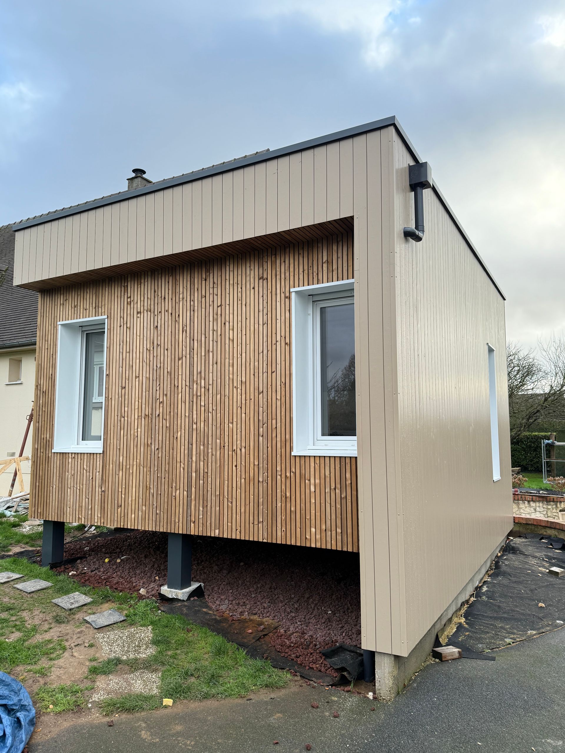 Maison moderne en bois avec bardage beige, fenêtres à cadre blanc, construite sur pilotis au-dessus d'un lit de gravier.