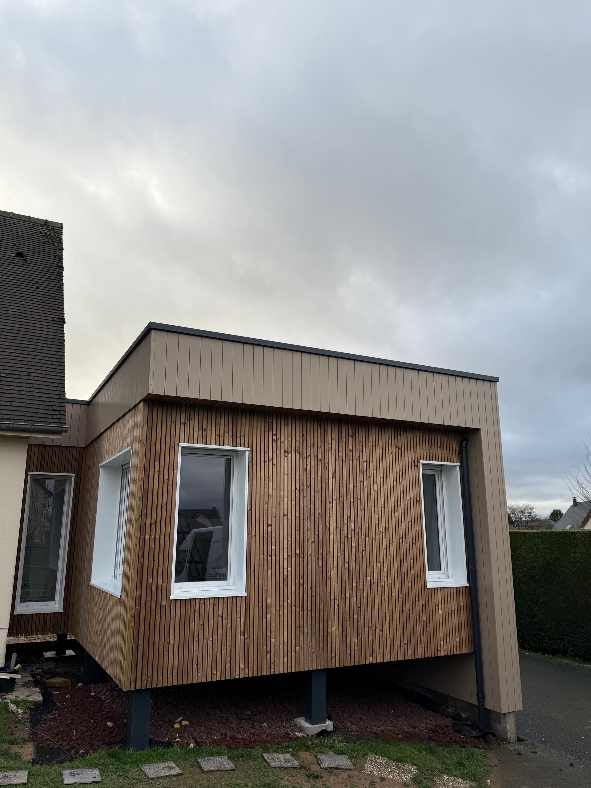 Extension moderne à panneaux de bois et fenêtres à cadre blanc, attenante à une maison. Ciel gris foncé.