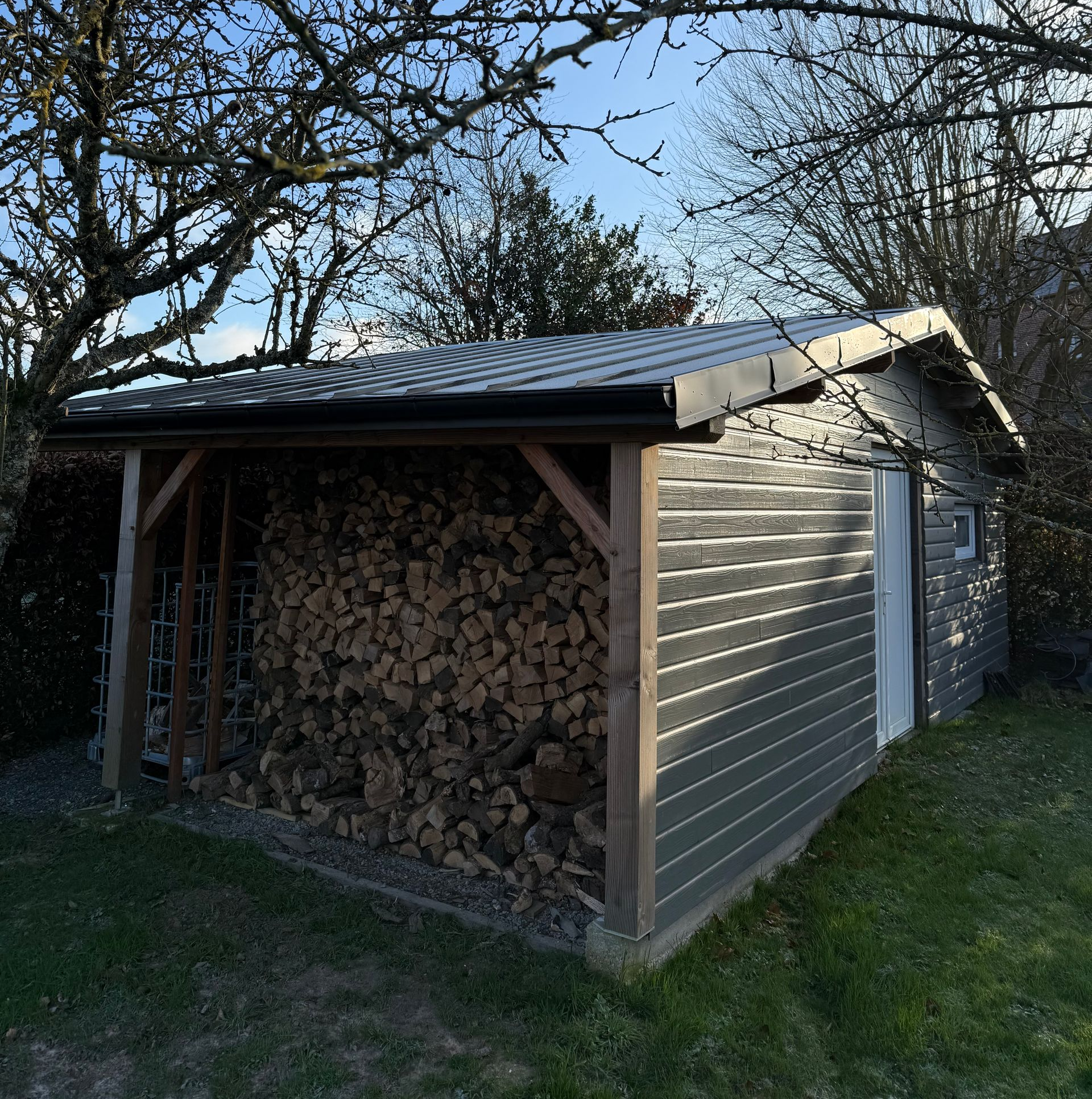 Abri à bois rempli de bois de chauffage, bardage gris, toit noir. Poutres de soutien en bois.