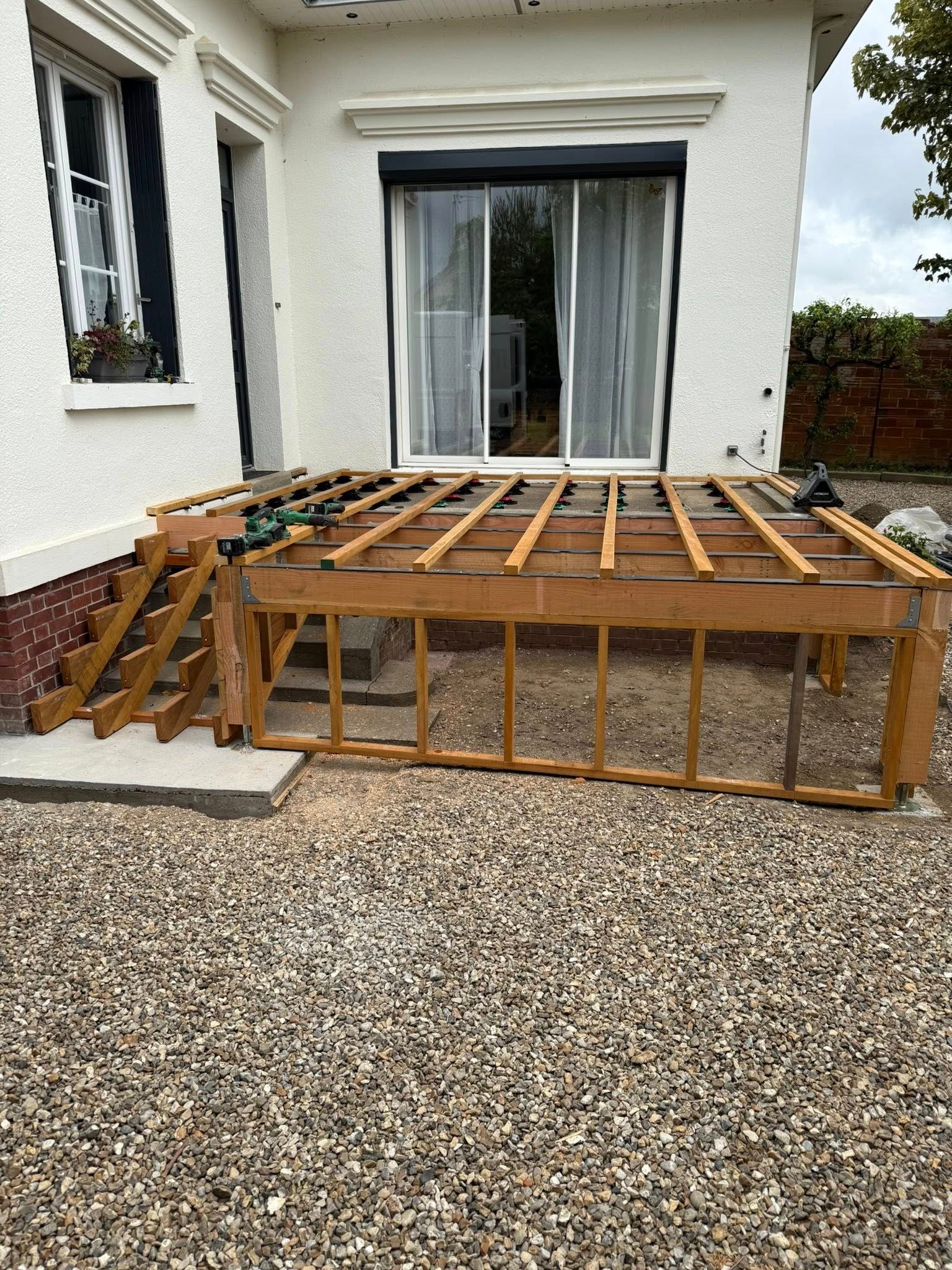 La structure d'une terrasse en bois est en construction à l'extérieur d'un bâtiment blanc. Sol en gravier.