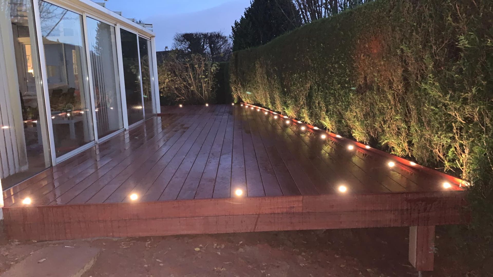 Terrasse en bois avec éclairage intégré, jouxtant une structure aux parois de verre et une haie illuminée. Ambiance nocturne.
