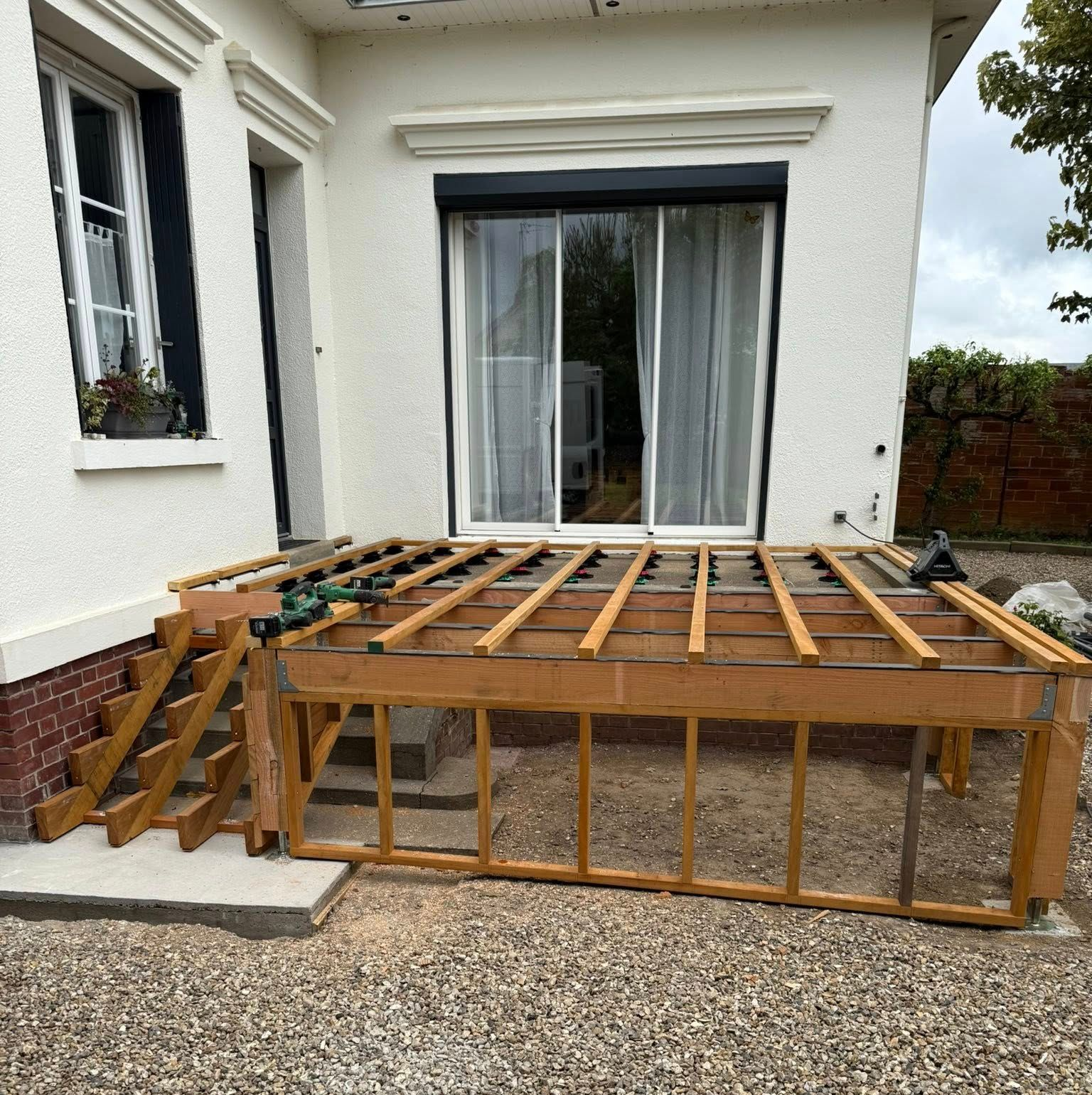 Terrasse en bois en construction à côté d'une maison avec porte, fenêtres et escalier.
