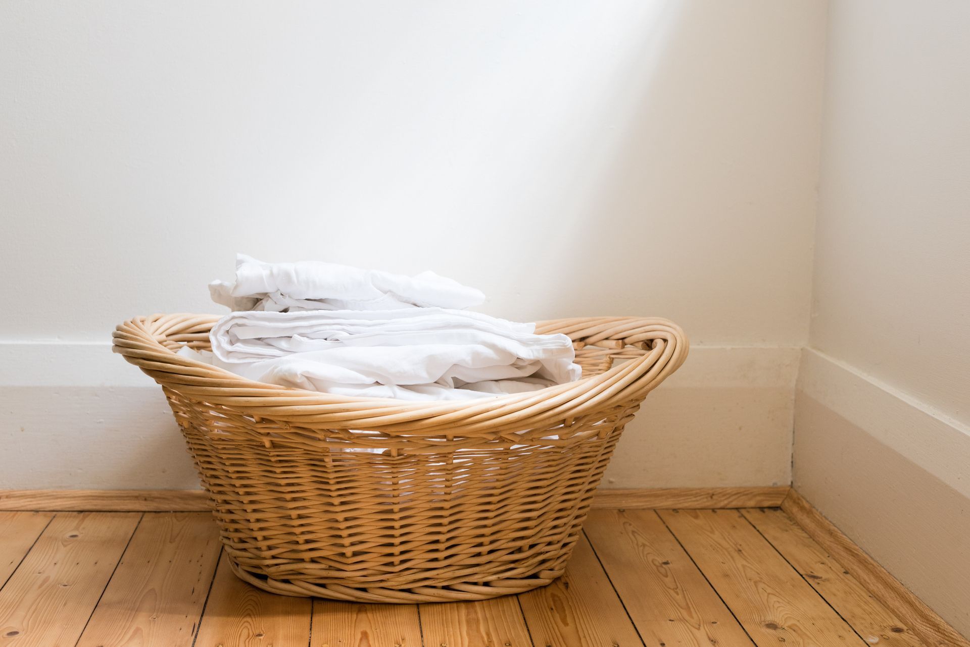 Panier en osier rempli de linge blanc plié, posé sur un parquet contre un mur blanc.