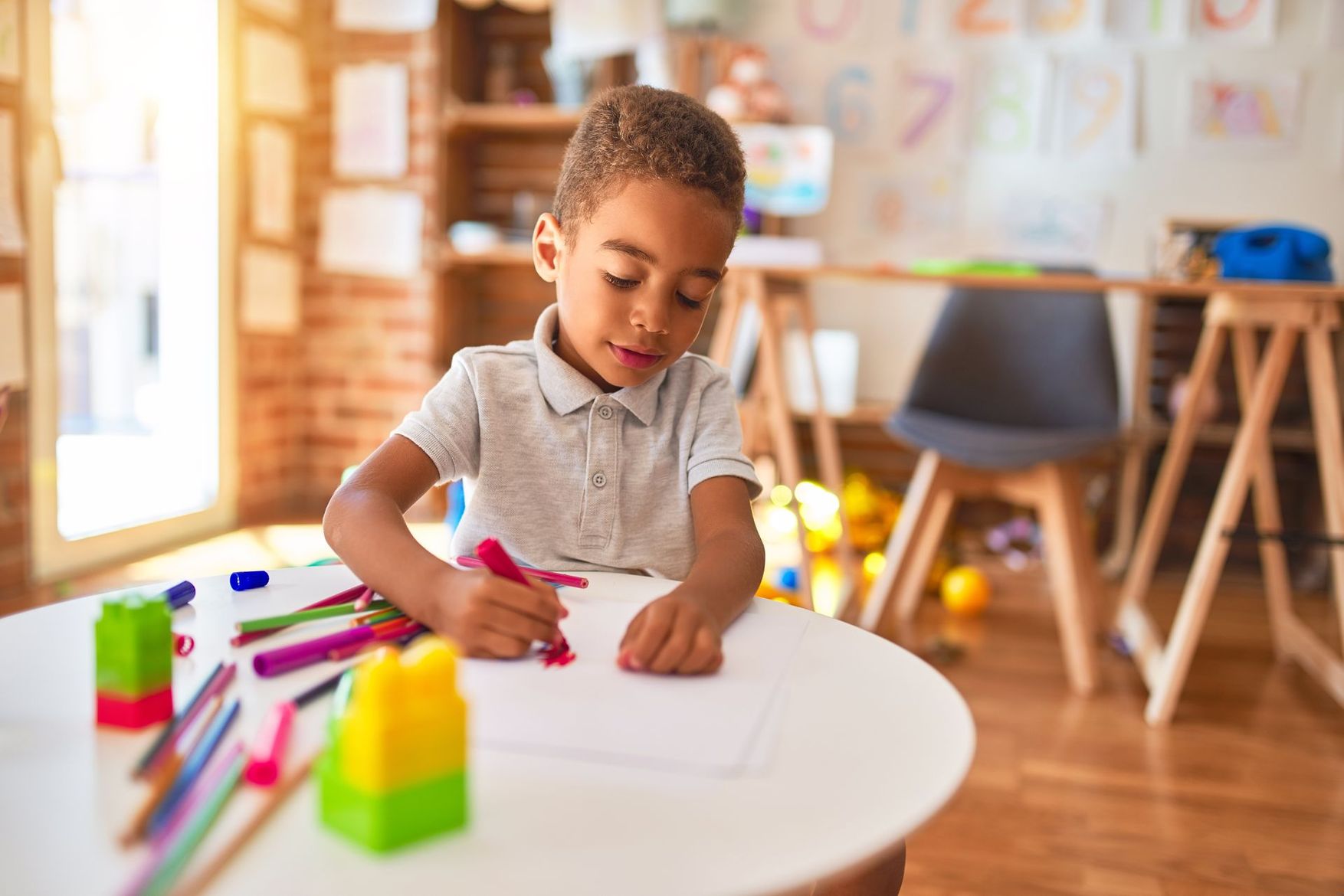 Un enfant colorie sur une table blanche dans une salle de classe lumineuse, avec des feutres et des blocs de couleur à proximité.