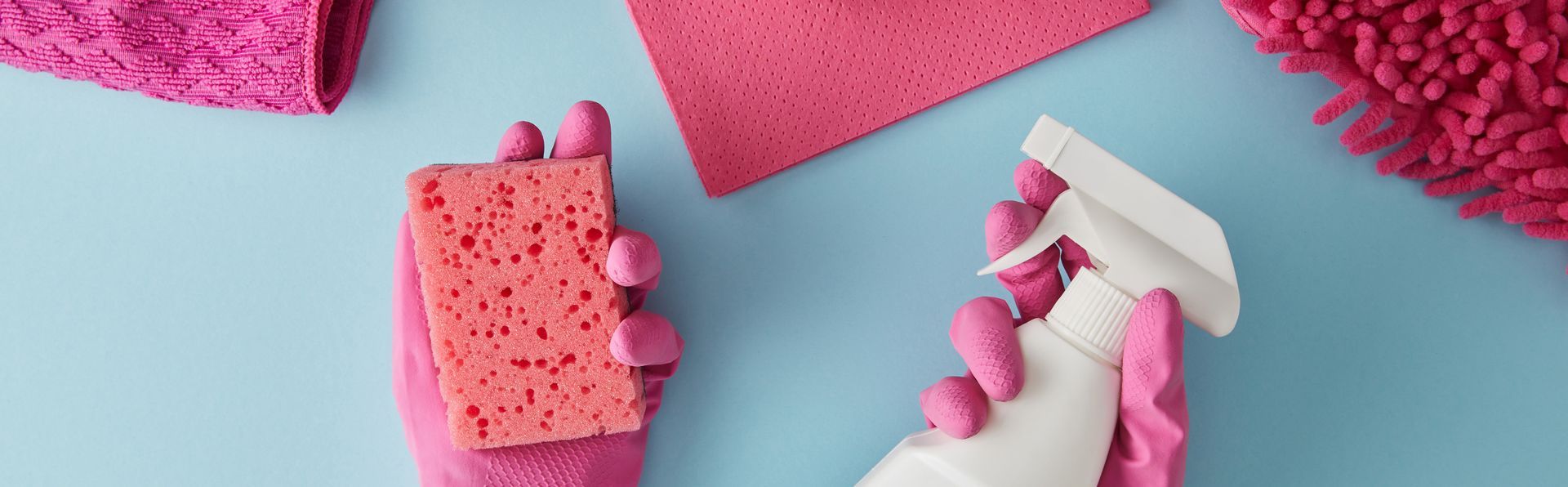 Outils de nettoyage roses et éponges sur fond bleu clair, une main tenant un vaporisateur