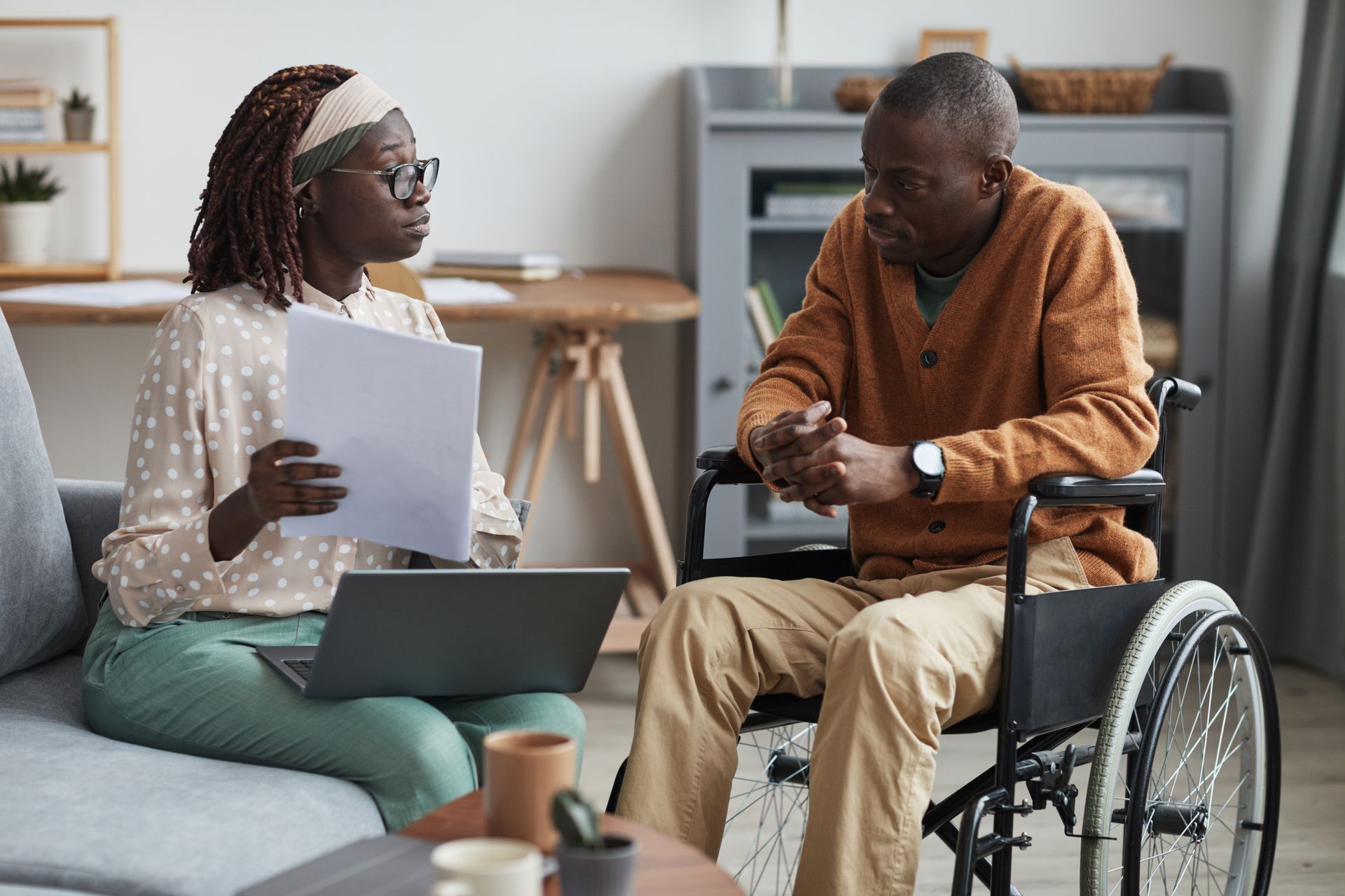 Deux personnes discutent de documents administratifs dans un salon, l'une d'elles est assise dans un fauteuil roulant avec un ordinateur portable ouvert.