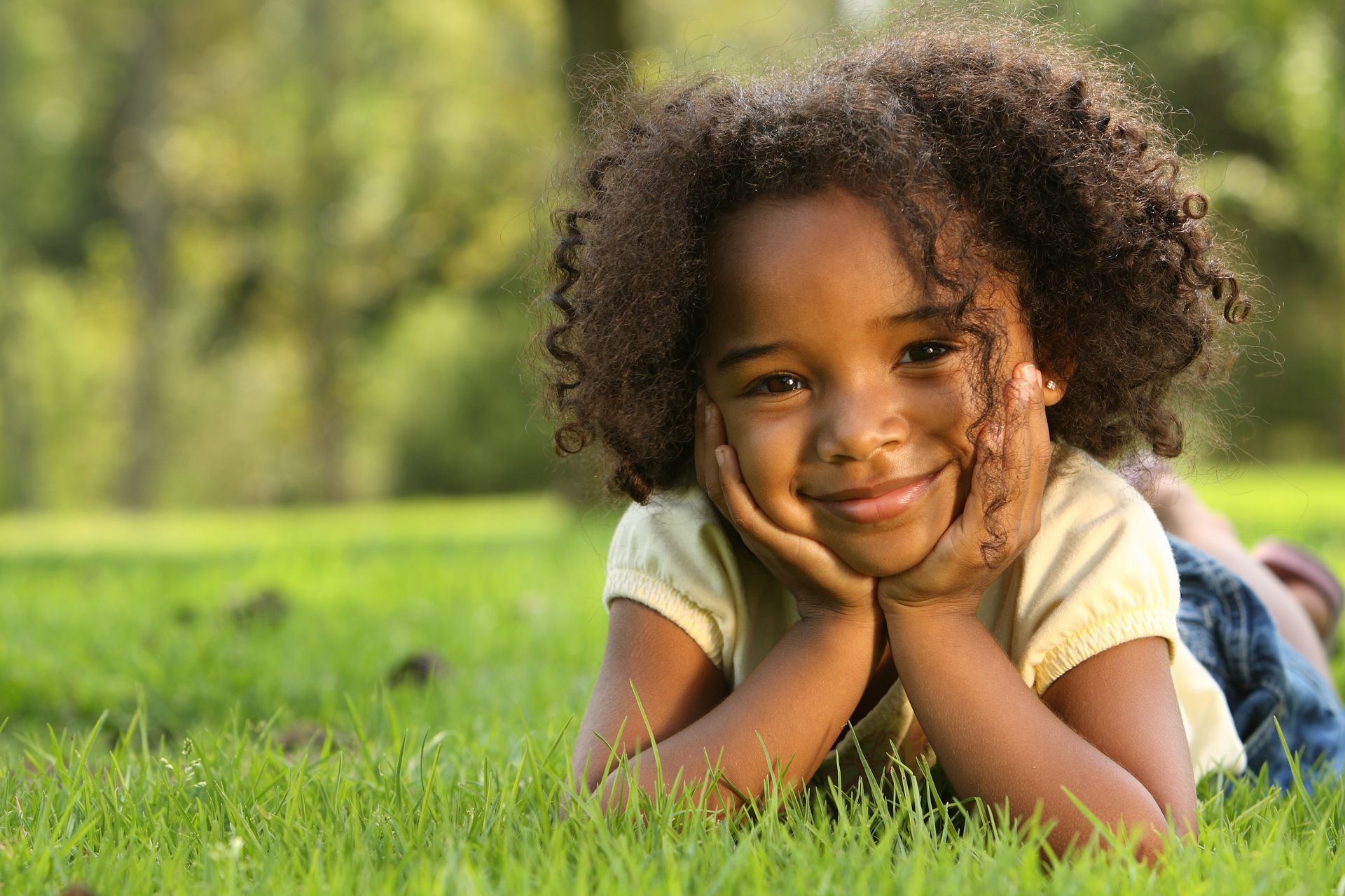 Enfant souriant allongé dans l'herbe, les mains sur les joues, dans un parc ensoleillé