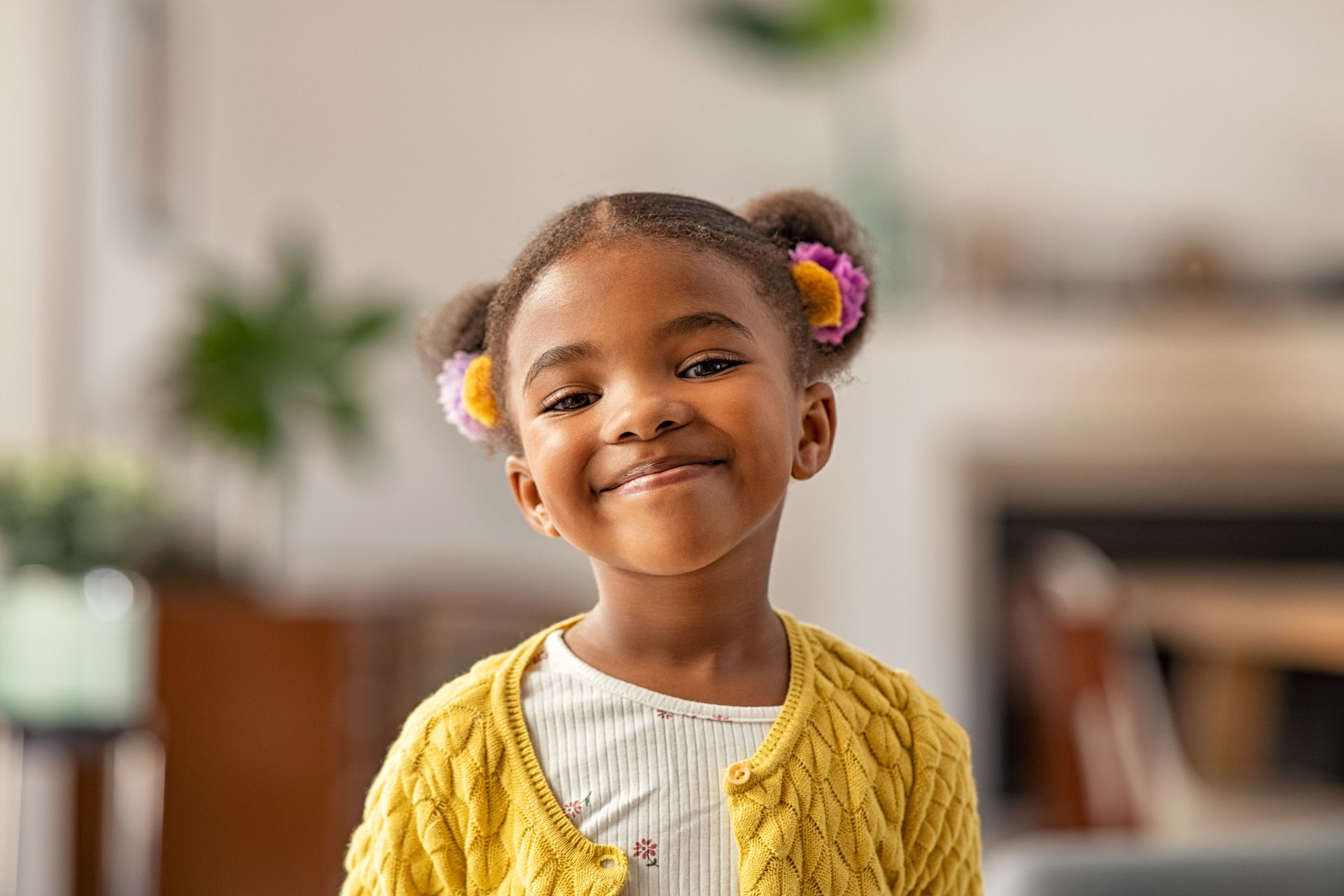 Enfant souriant avec des élastiques colorés dans les cheveux, portant un cardigan jaune, à l'intérieur avec un arrière-plan légèrement flou.