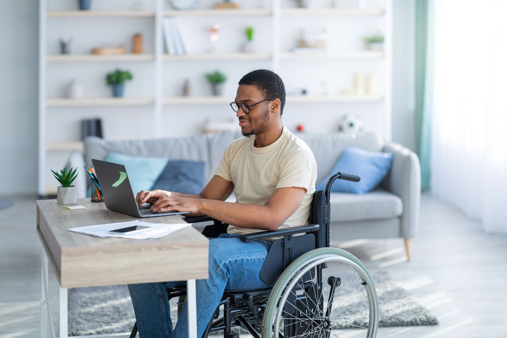 Un homme en fauteuil roulant utilise un ordinateur portable à un bureau dans un salon lumineux.