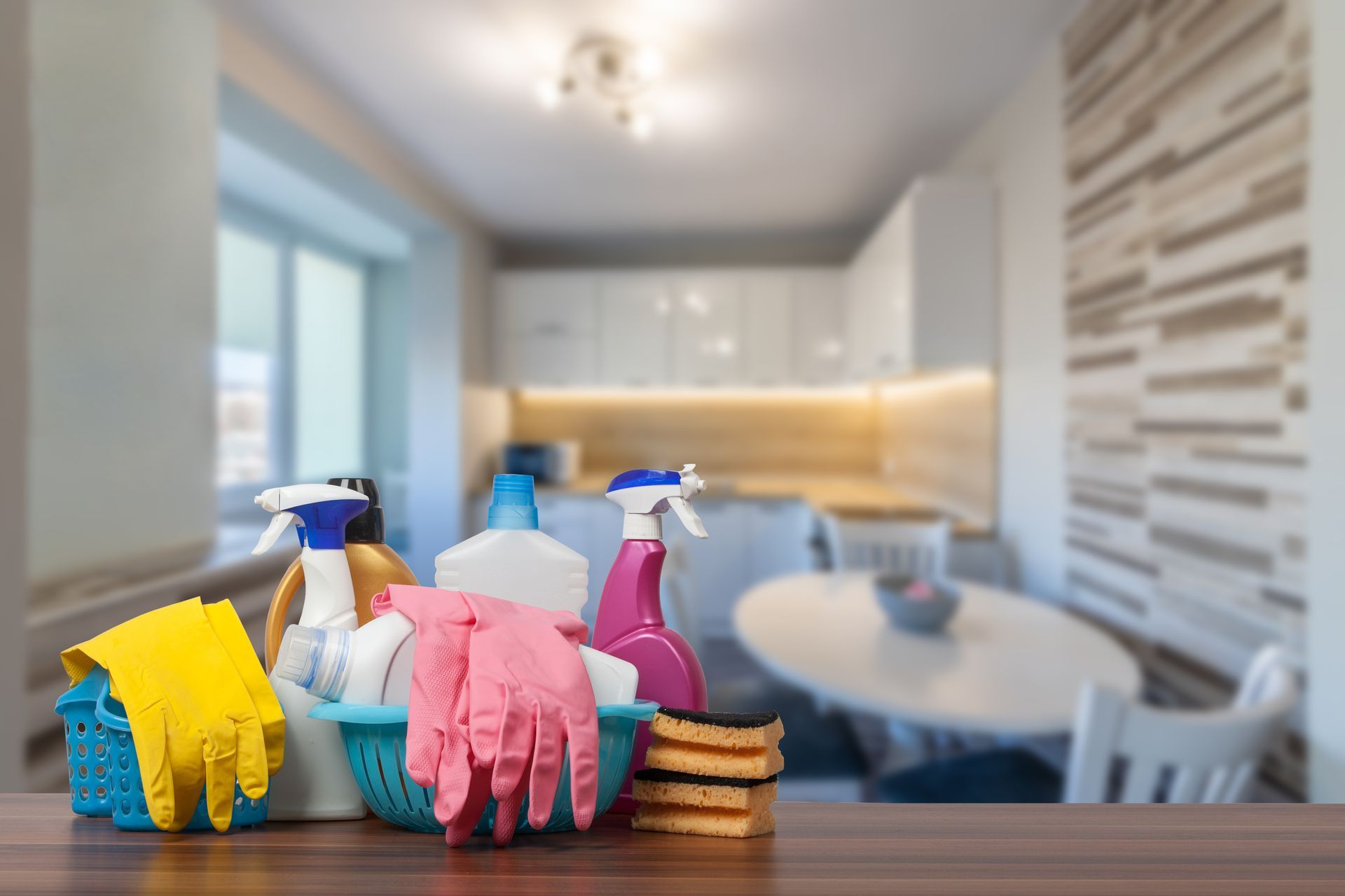 Des produits de nettoyage sont disposés sur une table dans une cuisine lumineuse : flacons pulvérisateurs, chiffons, gants et éponges.