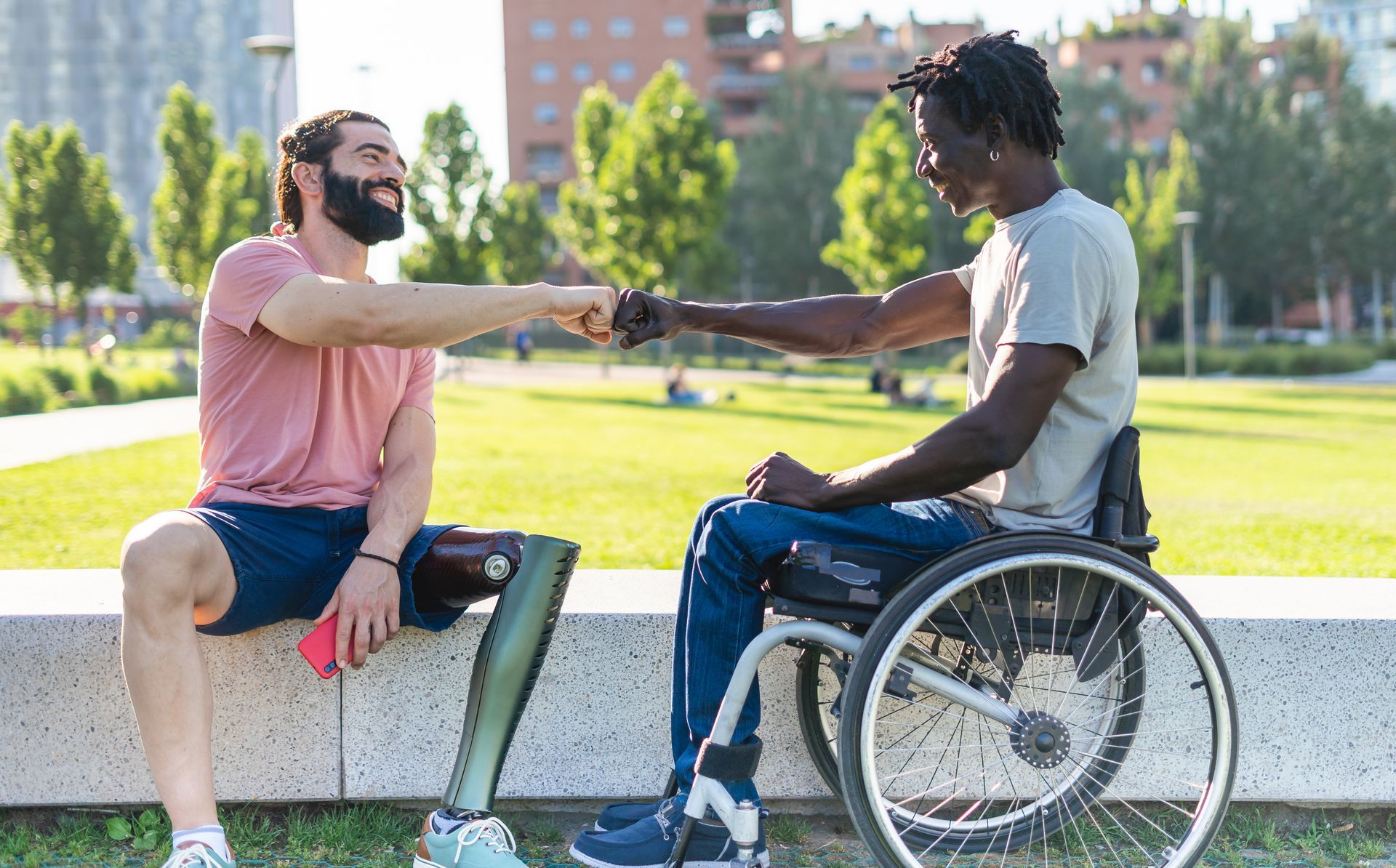 Deux personnes sont assises dehors, l'une sur un banc, l'autre en fauteuil roulant, souriant et se saluant d'un check.