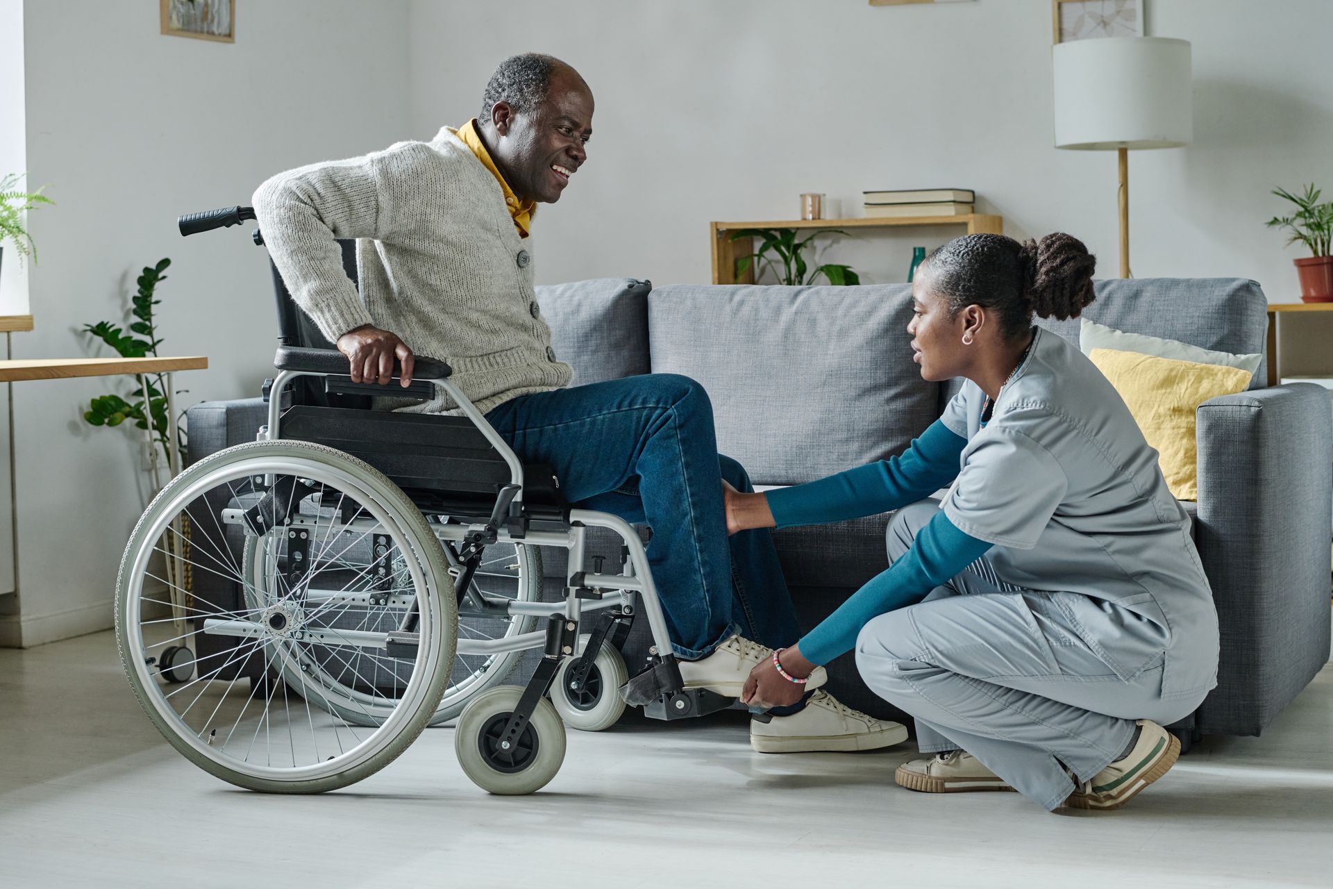 Une femme aide un homme à se transférer de son fauteuil roulant à un canapé dans un salon lumineux.