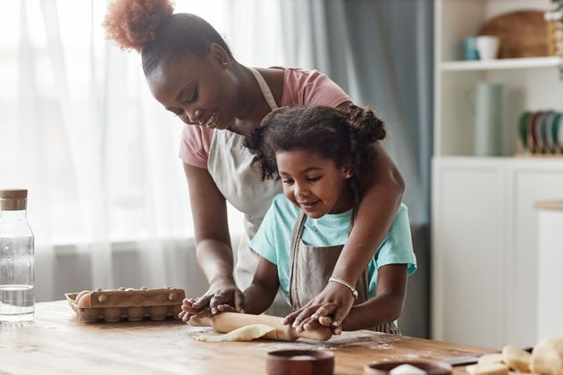 Un adulte aide un enfant à pétrir de la pâte à une table de cuisine près d'une fenêtre lumineuse.