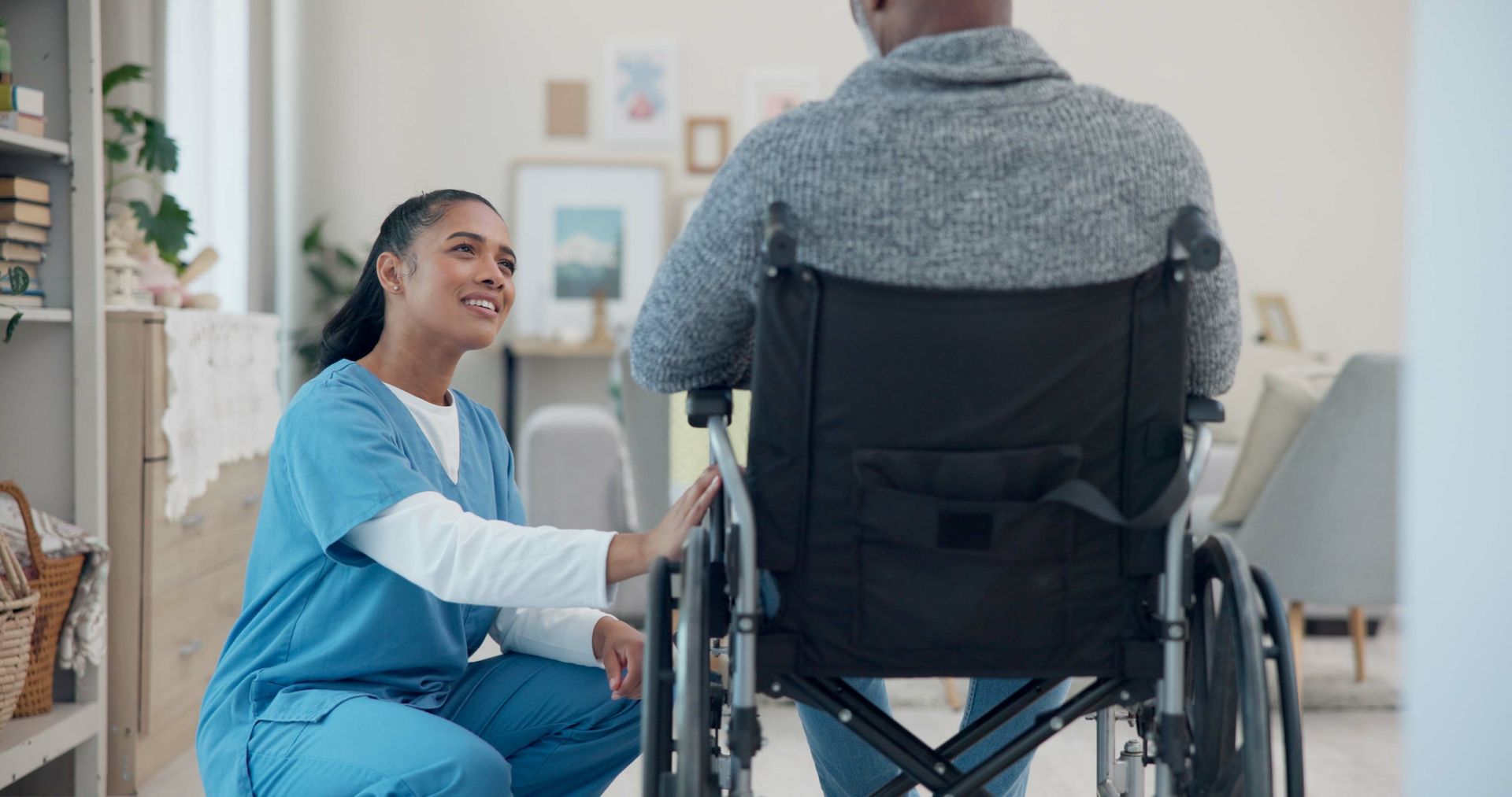 Une aide-soignante en blouse bleue sourit à côté d'une personne assise dans un fauteuil roulant à domicile