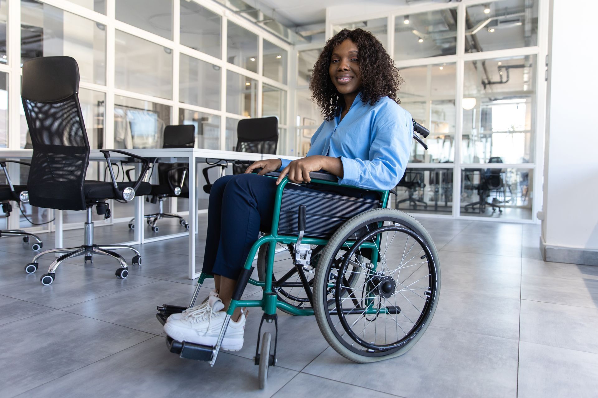Une femme en fauteuil roulant sourit dans un bureau moderne aux murs et bureaux en verre.