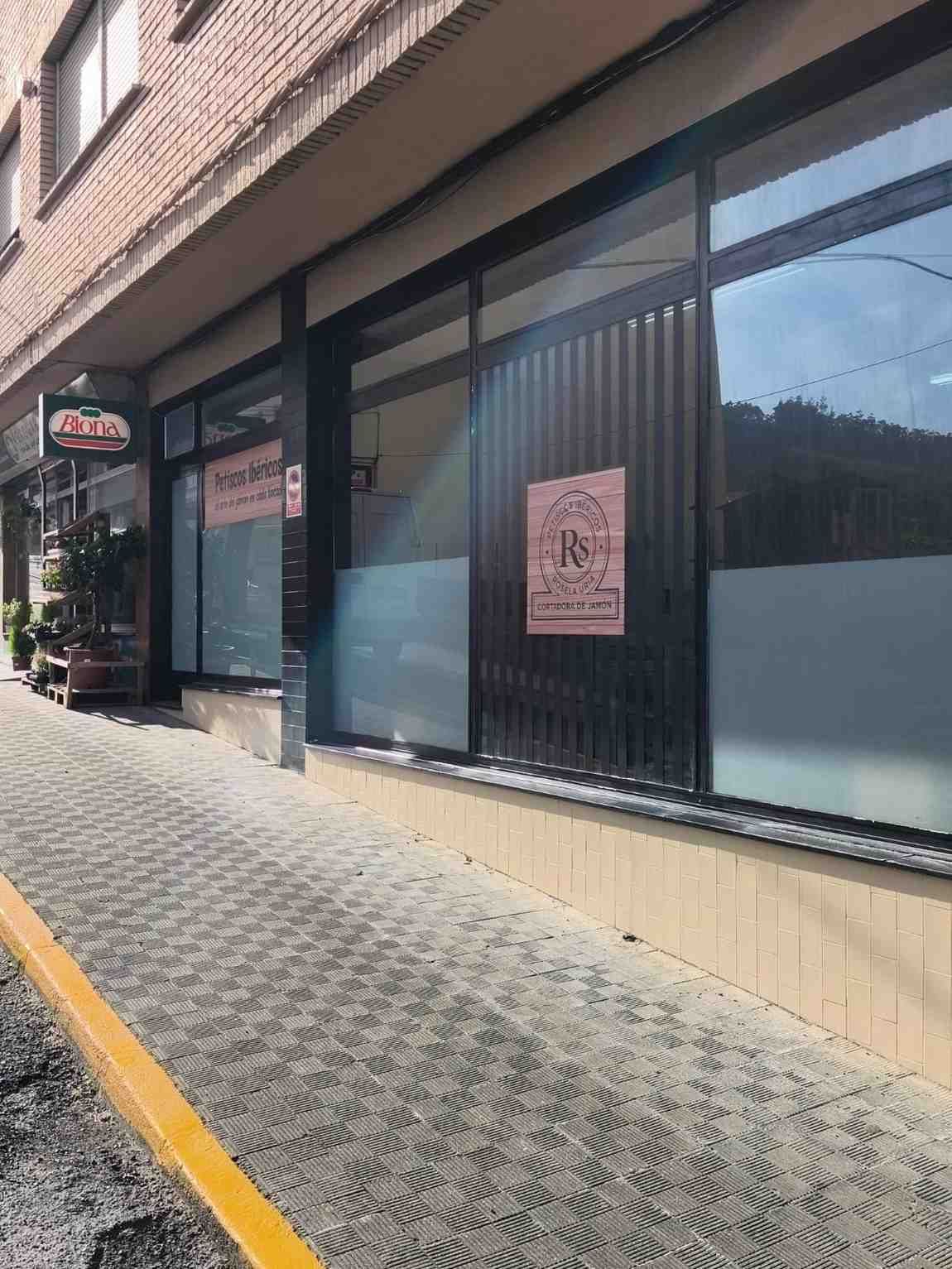 Exterior de un edificio con grandes ventanales y cartel, acera de ladrillo y bordillo amarillo.