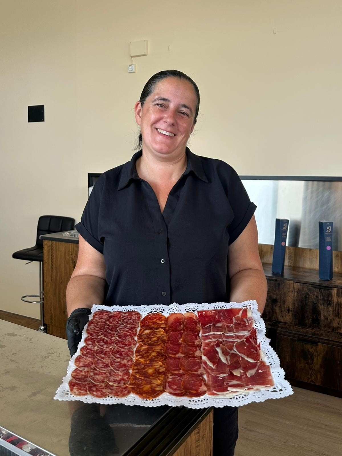 Mujer sonriendo, sosteniendo una bandeja de carnes cortadas. Camisa negra, ambiente de restaurante.