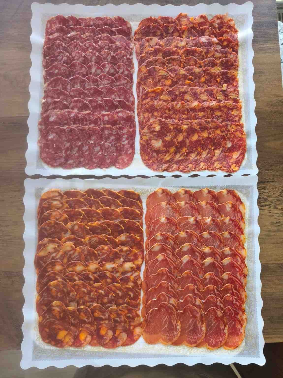 Cuatro bandejas de carnes cortadas en rodajas, de distintos tonos de rojo y naranja, dispuestas sobre una superficie de madera.