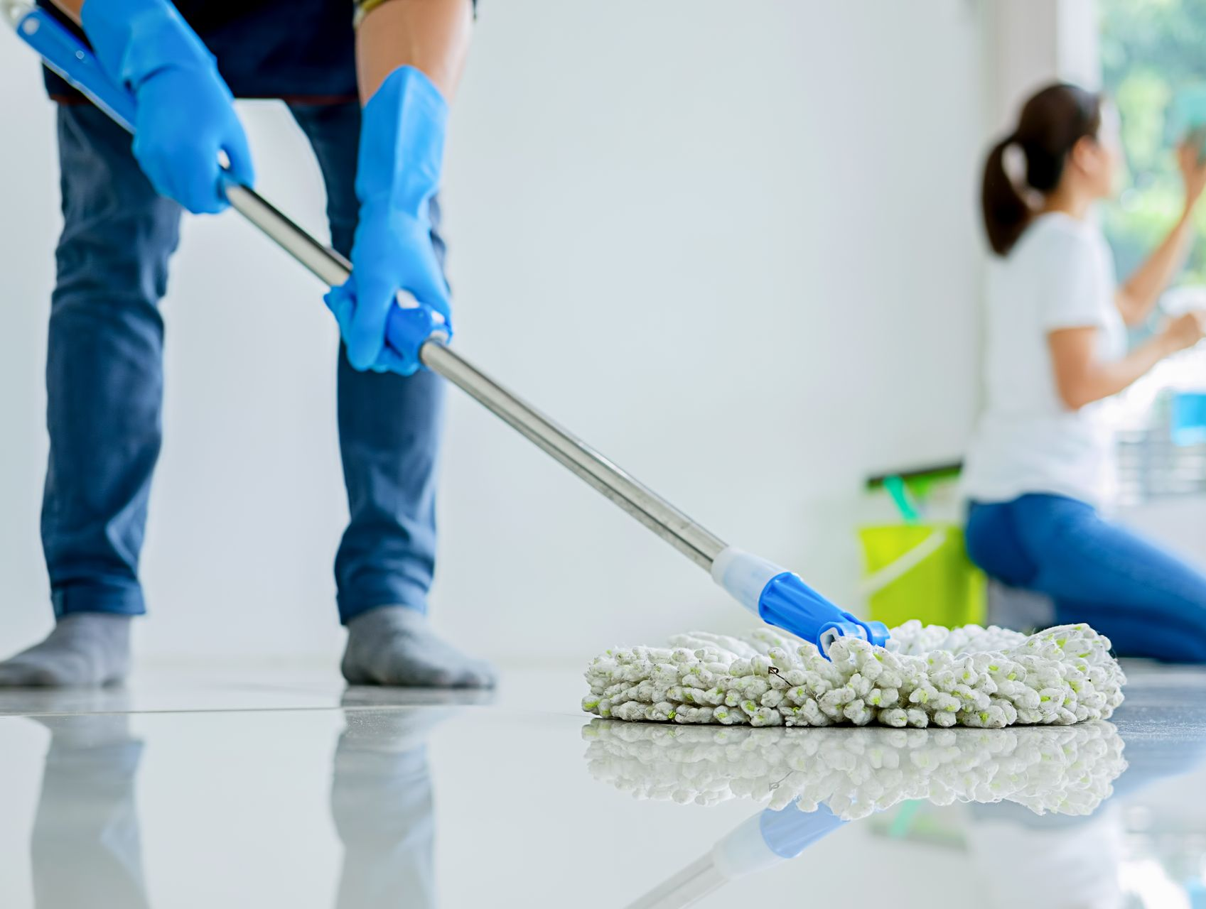 Une personne lave un sol blanc et brillant. Une autre personne, à l'arrière-plan, nettoie une vitre.