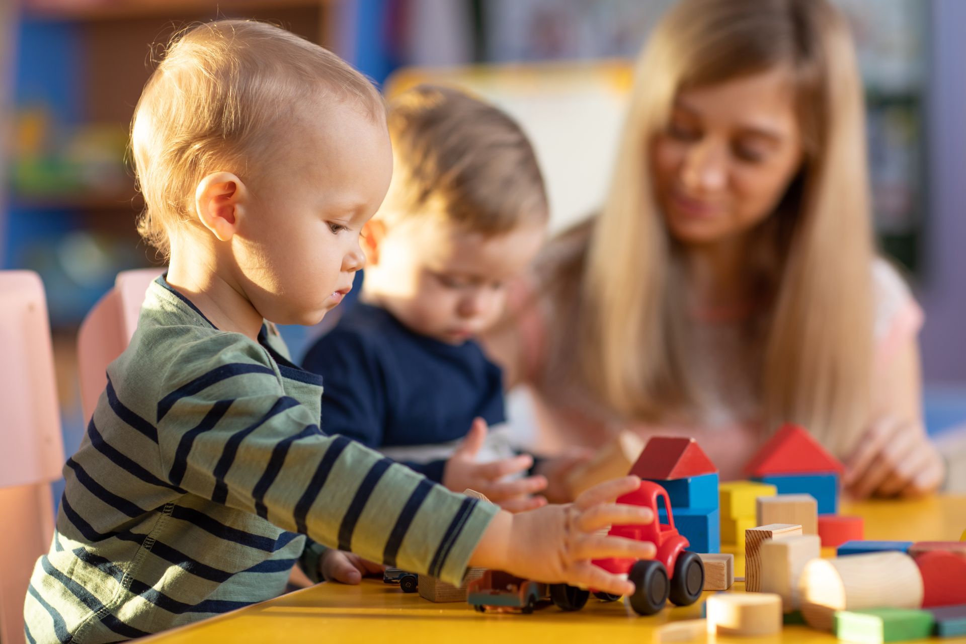 Un petit garçon joue avec un camion et des blocs de construction à une table, en présence d'un autre enfant et d'un adulte.