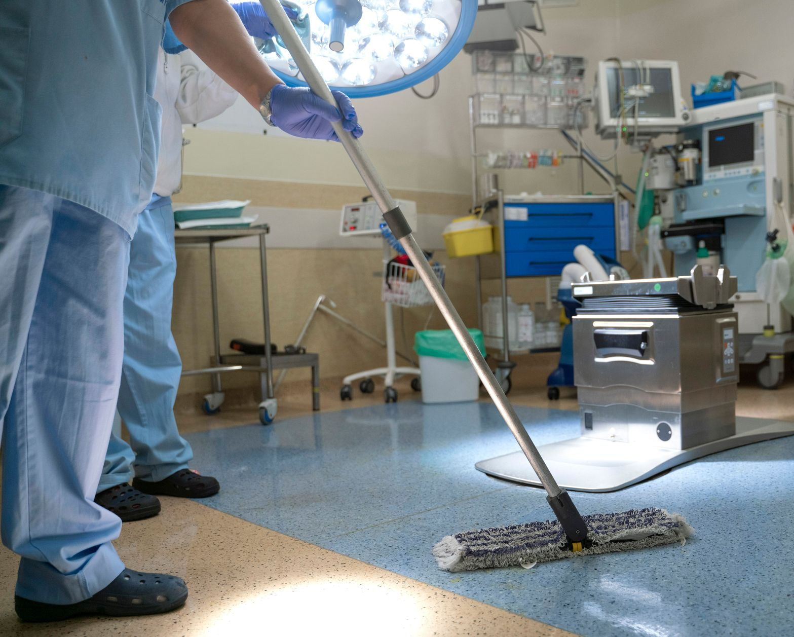 Une personne en blouse bleue nettoie le sol d'un hôpital bordé de lits, dans une palette de couleurs bleu clair et jaune.
