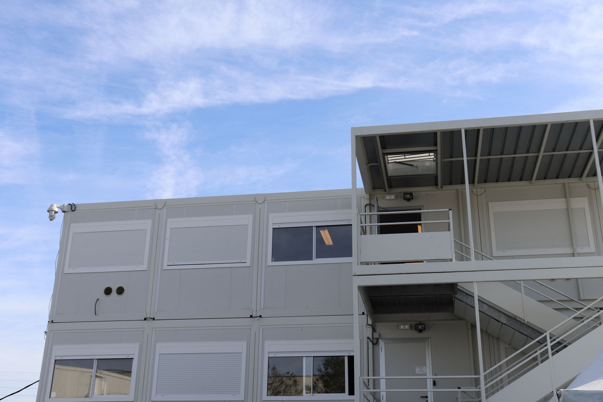 Bâtiment modulaire gris de deux étages avec stores, escalier et toit en métal, se détachant sur un ciel partiellement nuageux.