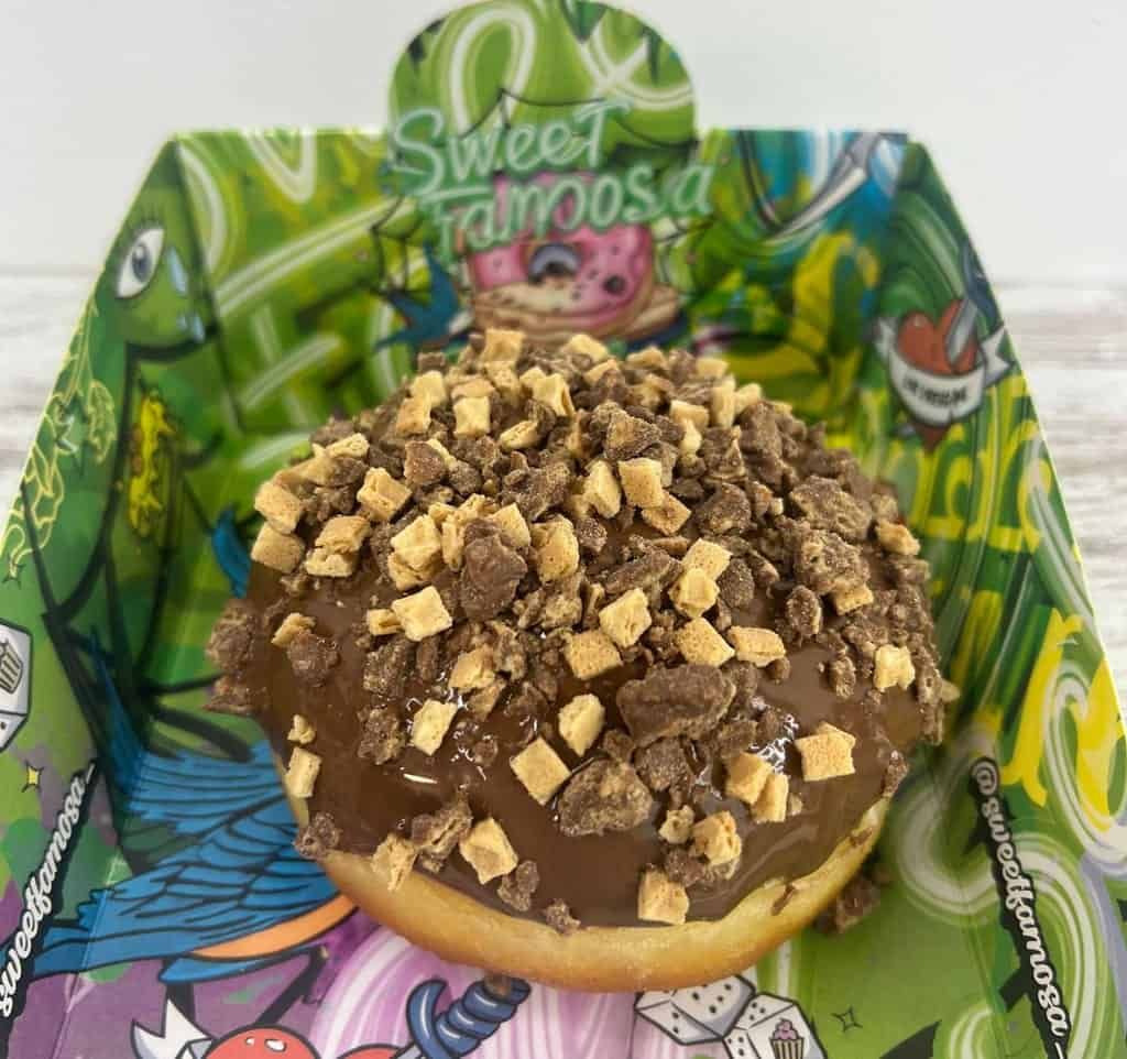 Una dona cubierta con glaseado de chocolate y cubierta con caramelos picados se encuentra en una colorida caja Sweet Yamosa con temática verde.