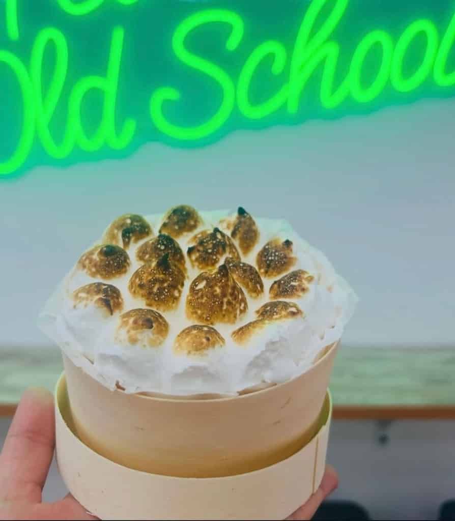 Una mano sostiene un pequeño recipiente color crema con un postre cubierto de merengue dorado. Un letrero de neón dice