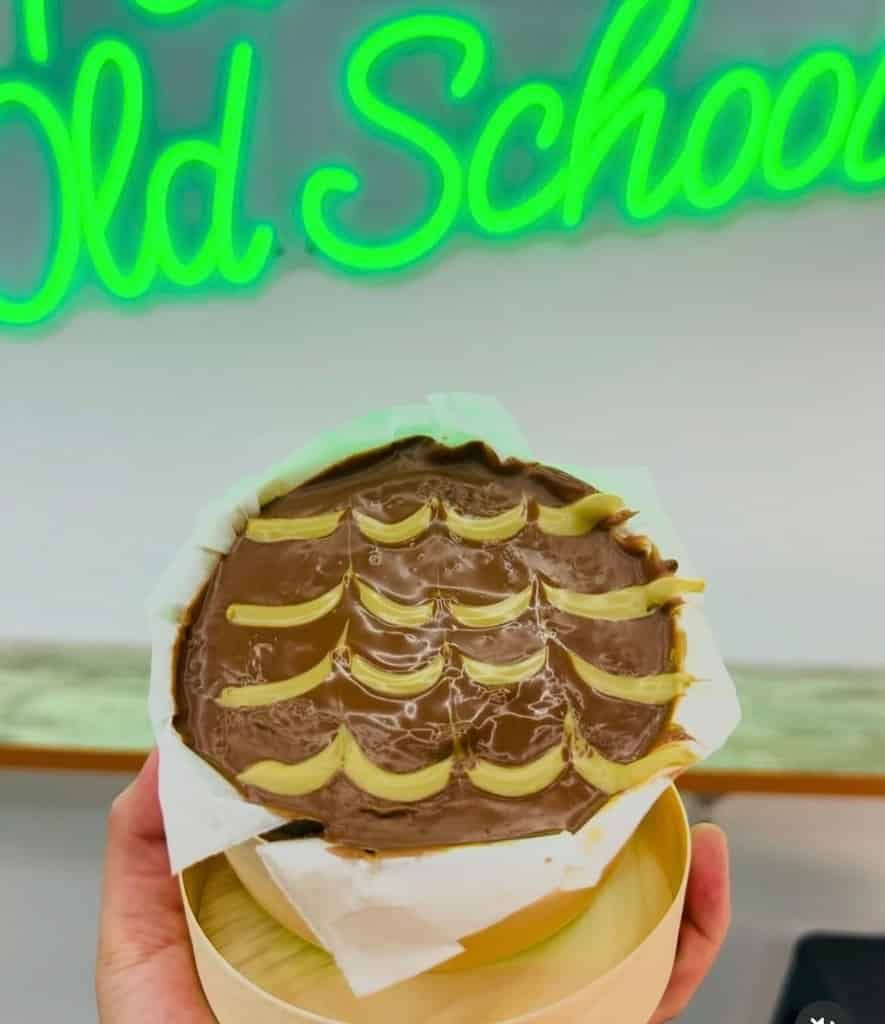 Una mano sostiene un postre redondo cubierto de chocolate con un patrón decorativo en un recipiente de madera, debajo de un letrero de neón verde