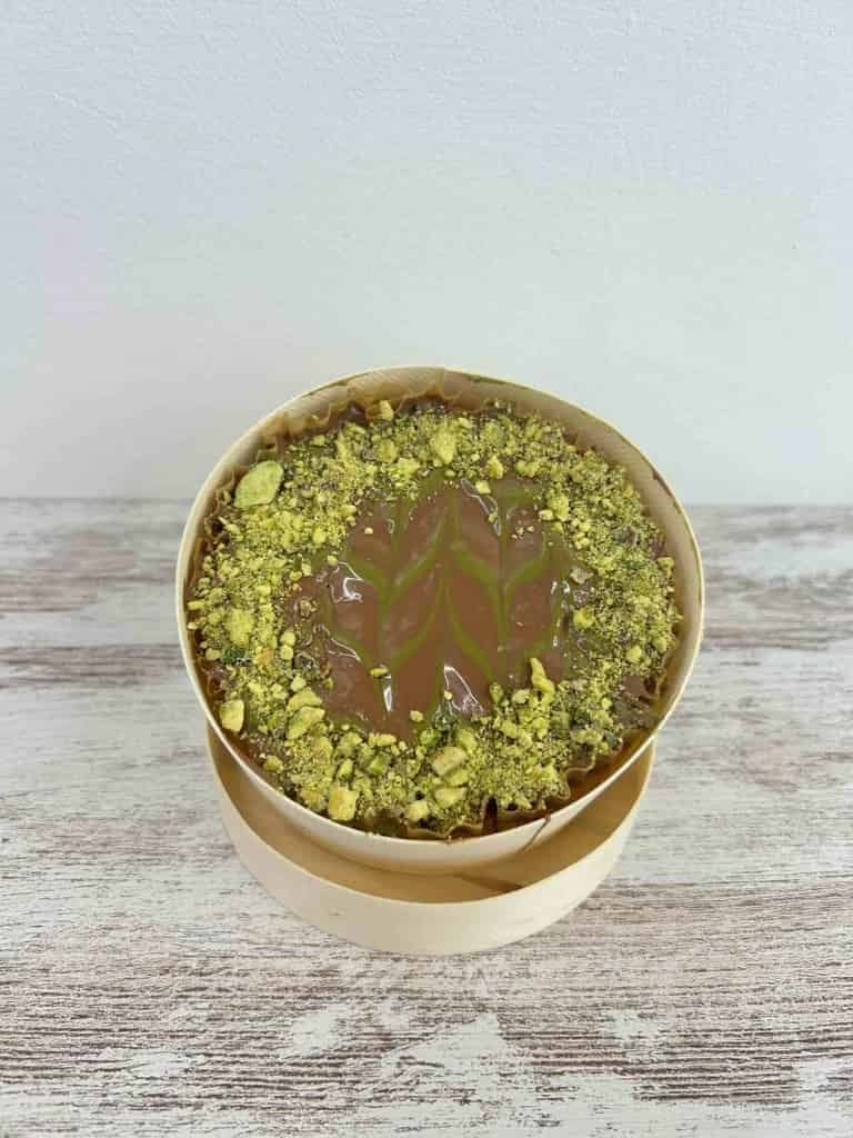 Un pequeño pastel redondo en una caja de madera, cubierto con pistachos verdes y un remolino de glaseado verde y marrón.