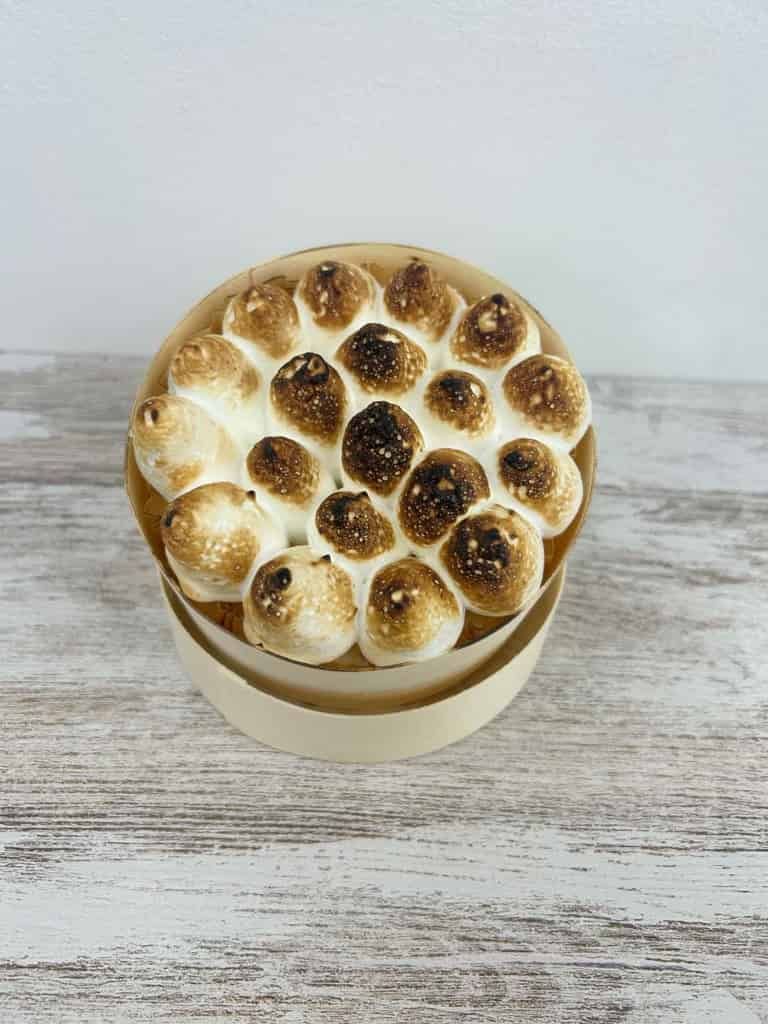 Un postre pequeño y redondo cubierto de malvaviscos tostados, dentro de un recipiente de color claro, reposa sobre una superficie de madera.