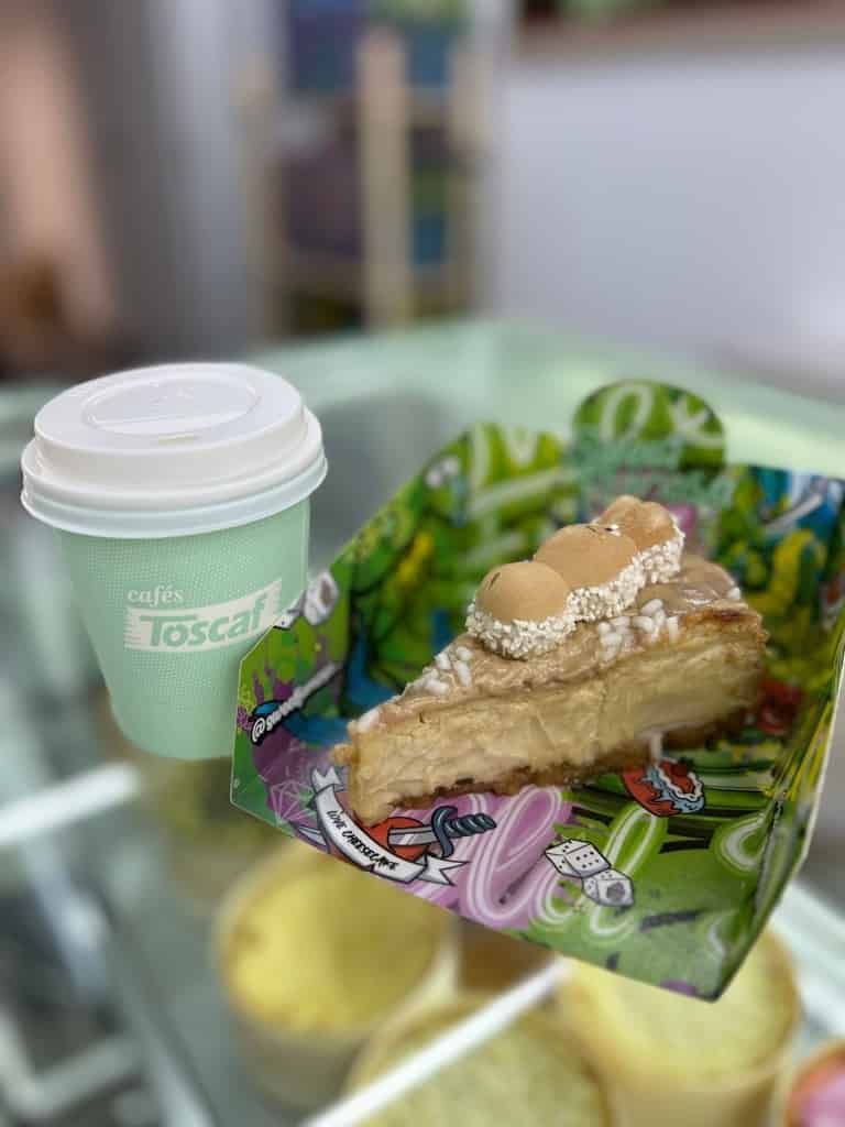 Una taza de café de color verde claro y una rebanada de tarta de queso con aderezos decorativos descansan sobre una bandeja de colores brillantes.