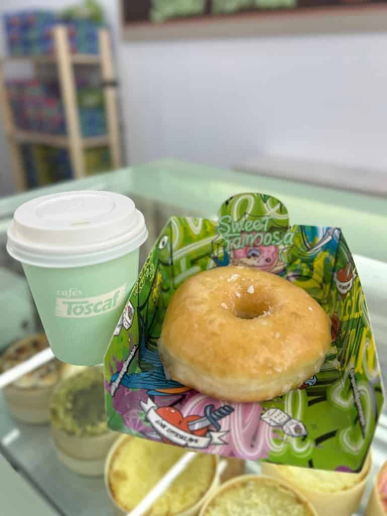 Donut en una caja colorida y una taza de café sobre un mostrador de vidrio en un entorno comercial.