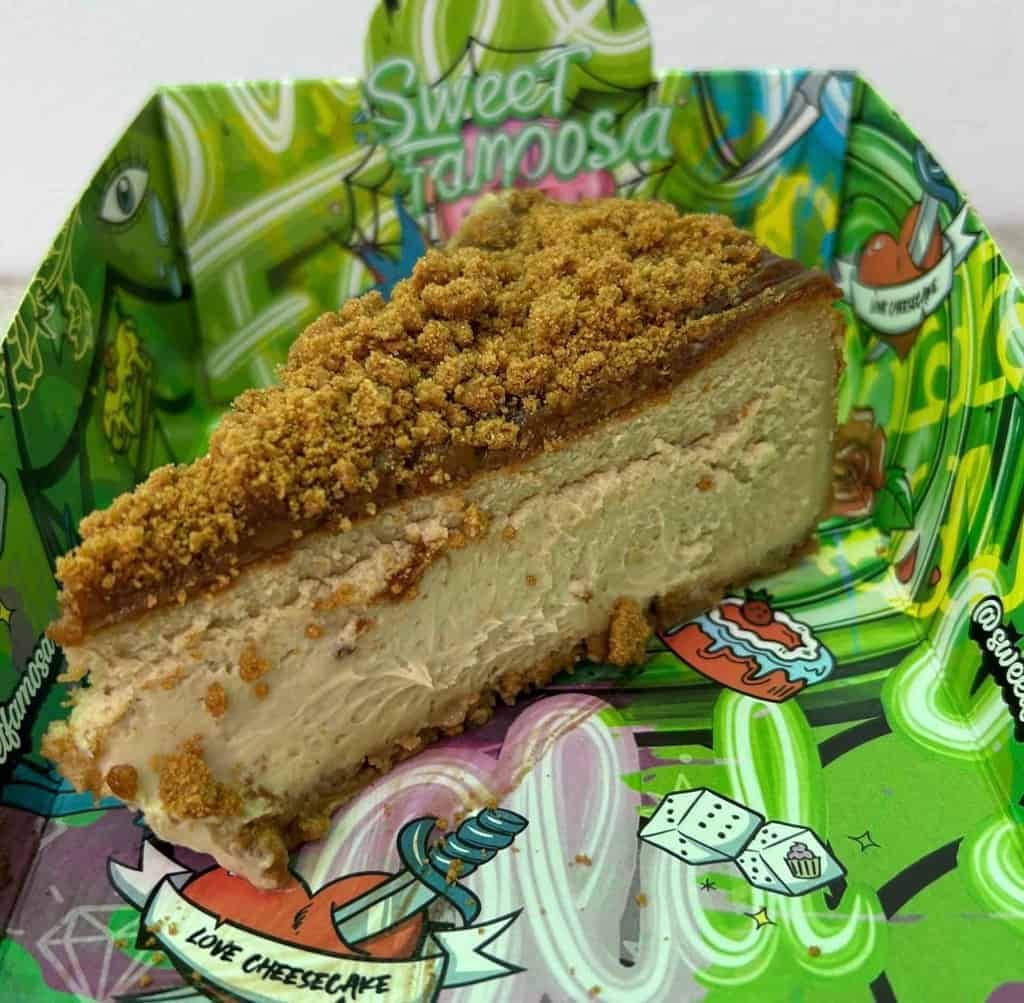 Una rebanada de tarta de queso cubierta con migas se encuentra en un recipiente de papel colorido con el texto