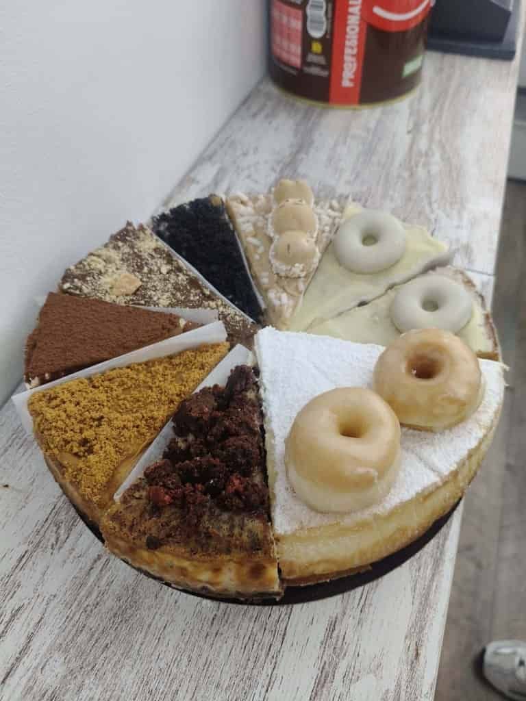 Una tarta con porciones de varios sabores, algunas de ellas con mini donas. Se ve un frasco de producto al fondo.