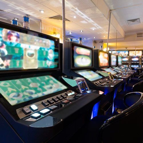 Fila de máquinas tragamonedas en un casino bien iluminado, pantallas que muestran gráficos de juegos coloridos, con sillas al frente.