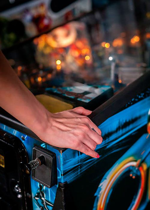 Mano apoyada en el lateral de una máquina de pinball azul; interior borroso con luces.