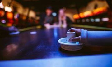 Mano de una persona sosteniendo un disco blanco sobre una mesa de hockey de aire; personas borrosas y escenario en el fondo.
