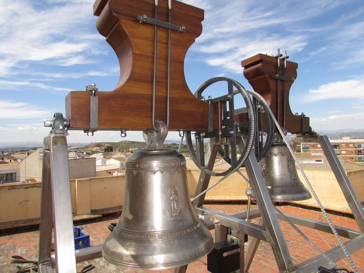 Dos grandes campanas de iglesia en lo alto de una torre, montadas sobre un marco de madera, con cielo azul de fondo.