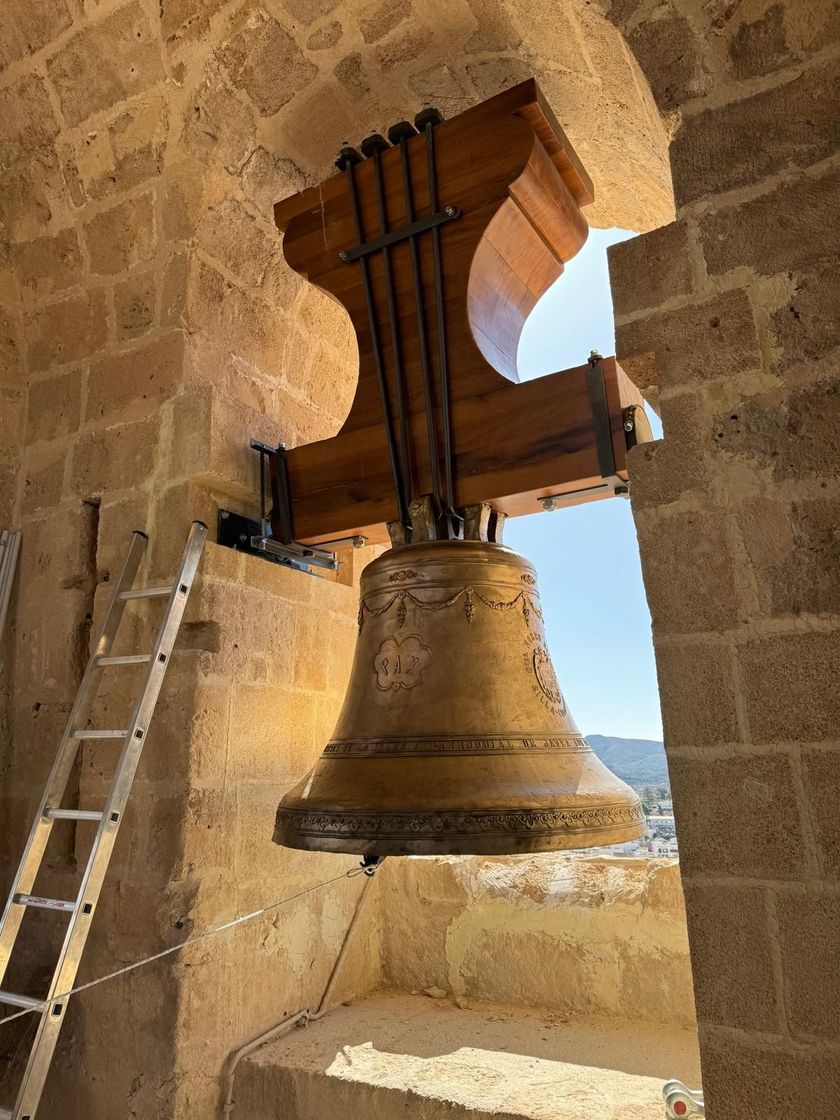 Una gran campana de bronce cuelga de un campanario de piedra, con una escalera apoyada contra la pared.